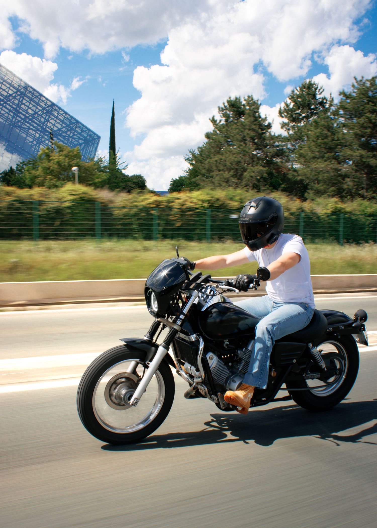 Person riding a black cafe racer motorcycle on a highway, wearing a helmet, white shirt, and jeans on a sunny day.