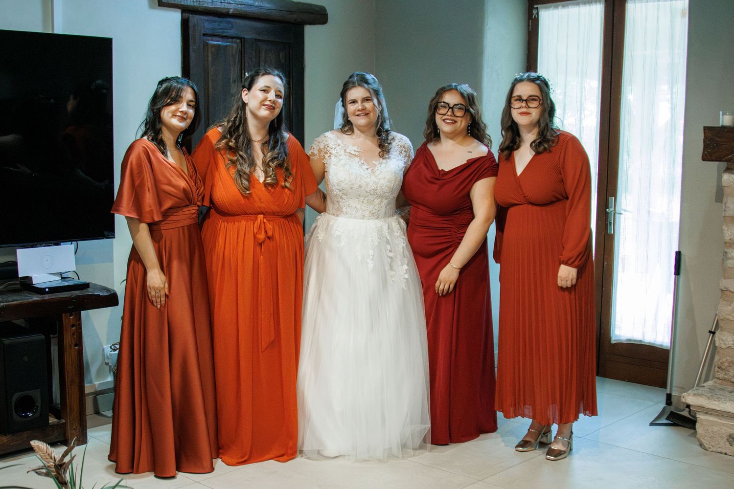 Bride in white lace gown poses with four bridesmaids in rust and red dresses indoors at a wedding.