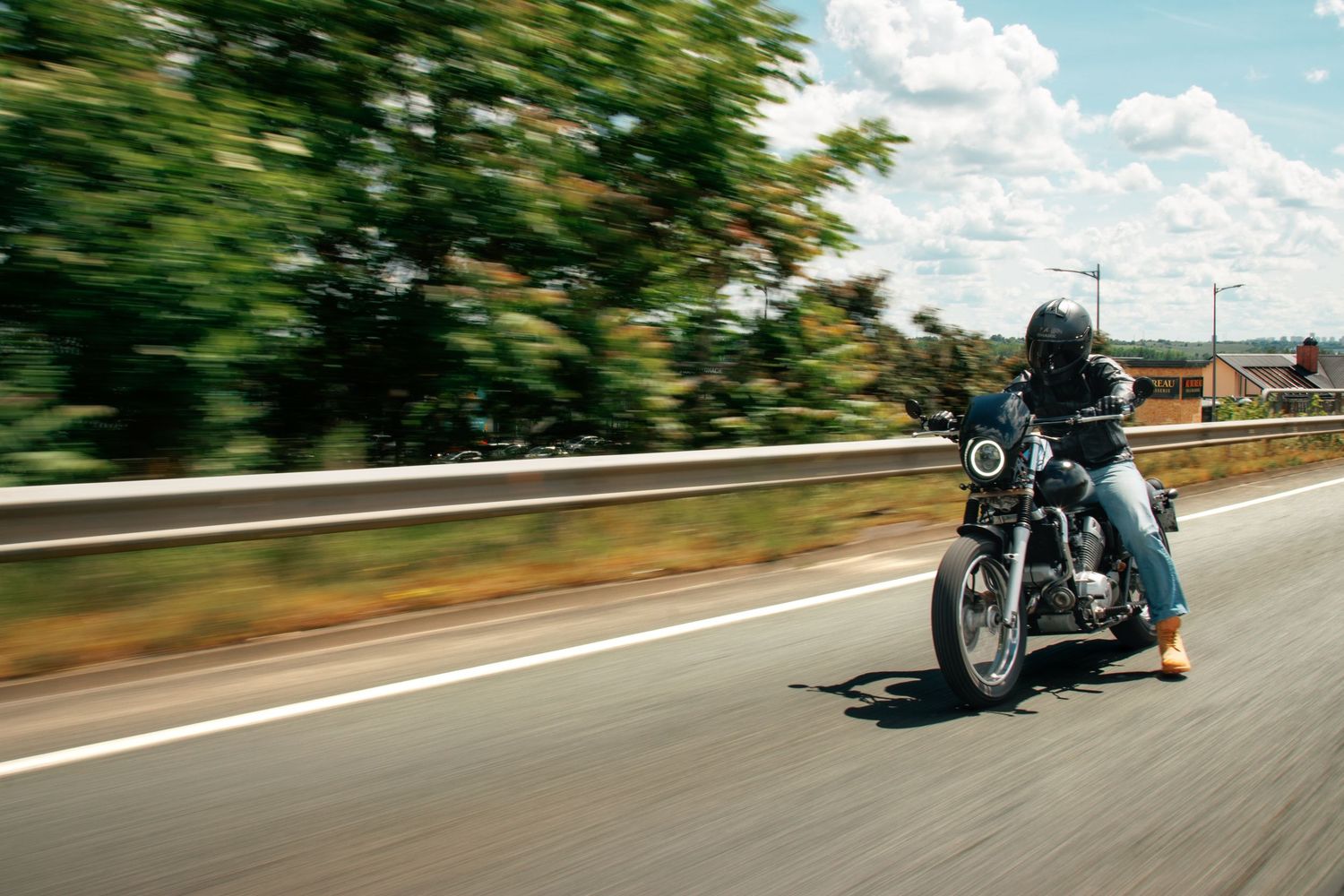 Motorcyclist riding a classic cruiser motorcycle at high speed on a highway surrounded by lush green trees.