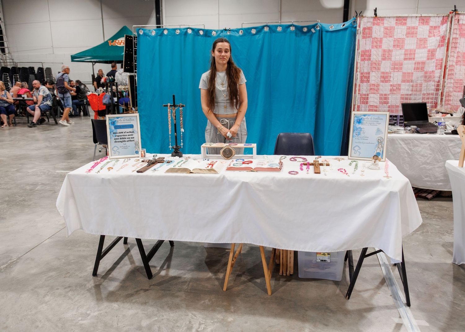Woman standing behind a white draped vendor table at an indoor craft fair with blue backdrop.