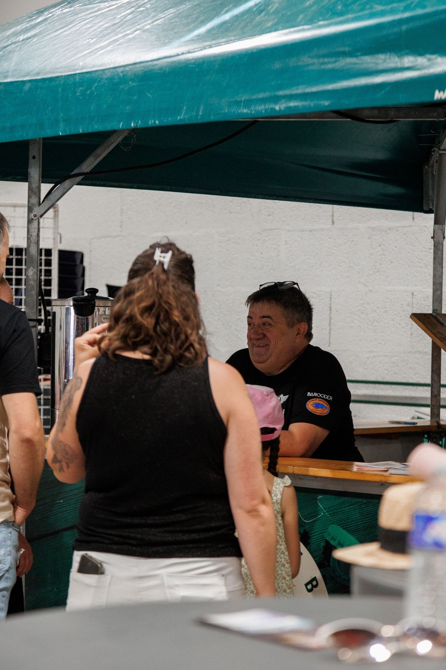 People gathered under a teal canopy tent at an outdoor event, with a smiling man in a black shirt conversing with others.