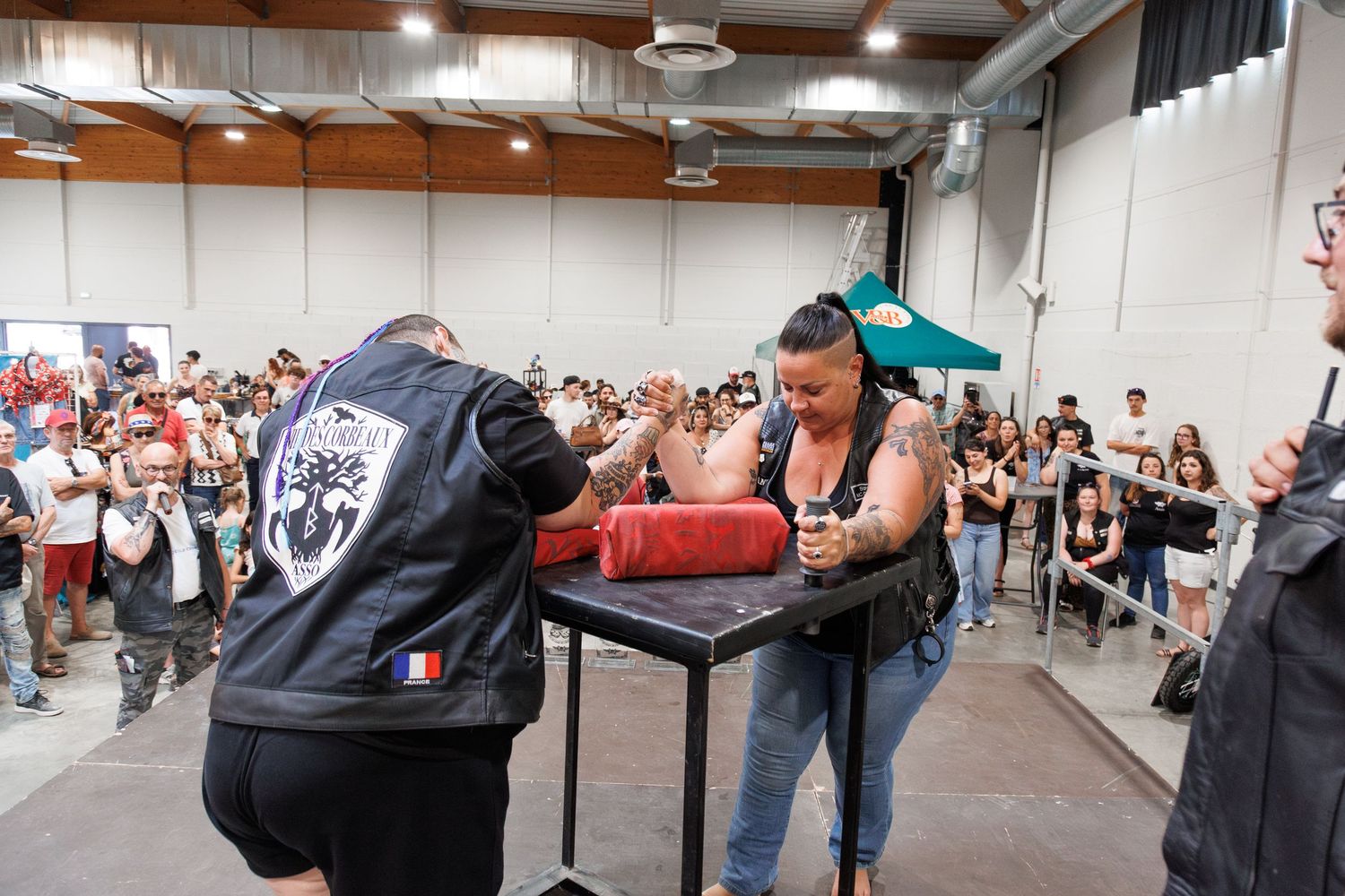 Two competitors arm wrestling at a public event in a crowded indoor venue, with spectators watching in the background.