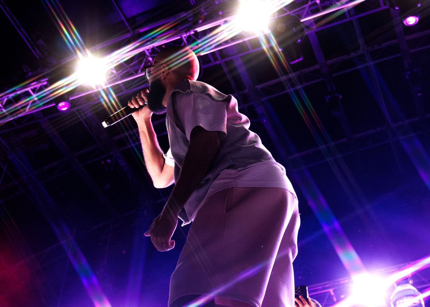 A performer silhouetted against dramatic purple stage lights holds a microphone while singing during a concert performance.