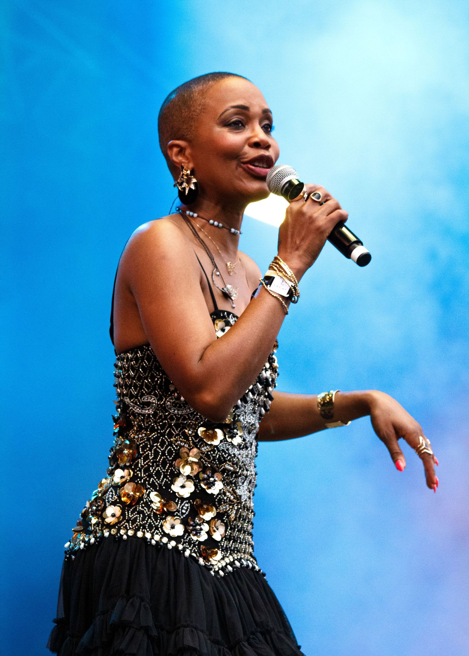 A performer in a black and silver embellished dress sings passionately against a bright blue sky backdrop during an outdoor show.