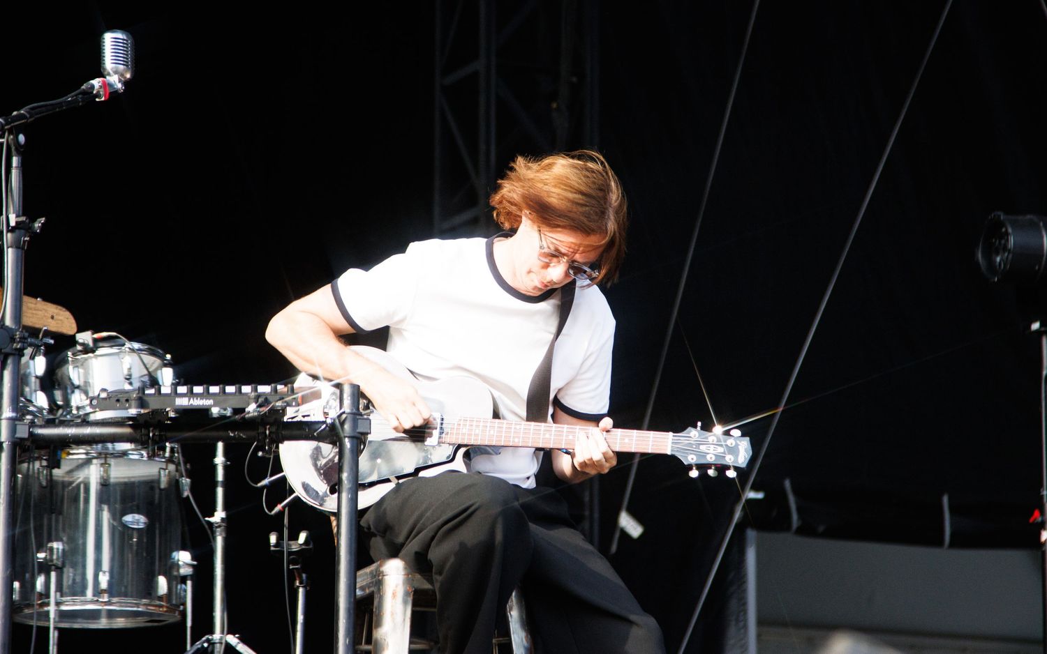 A young guitarist with red hair performs on an outdoor stage, playing a white electric guitar during a live concert.