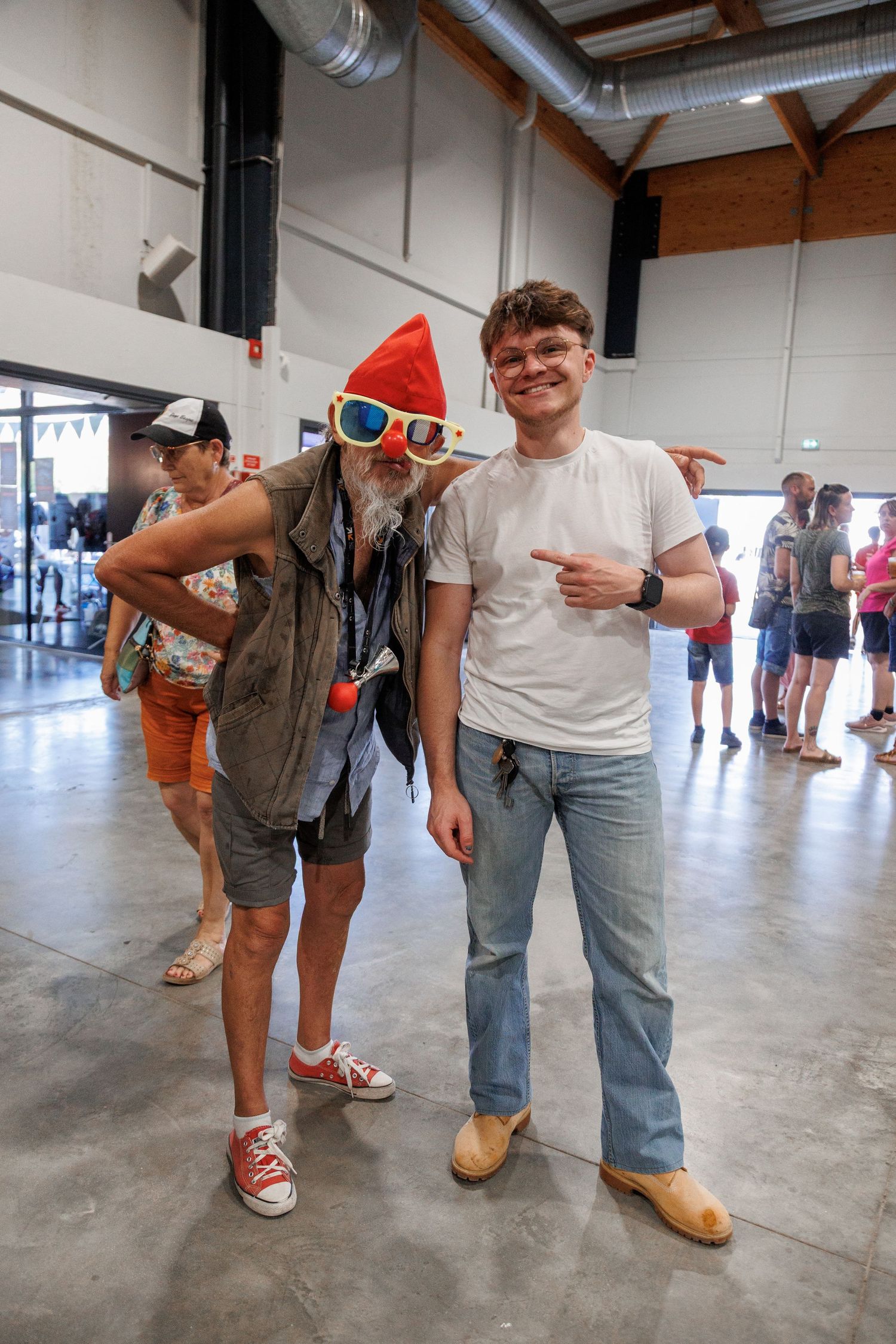 Two men pose together at an indoor event, one wearing a red gnome hat and oversized sunglasses, the other in jeans and a white tee.