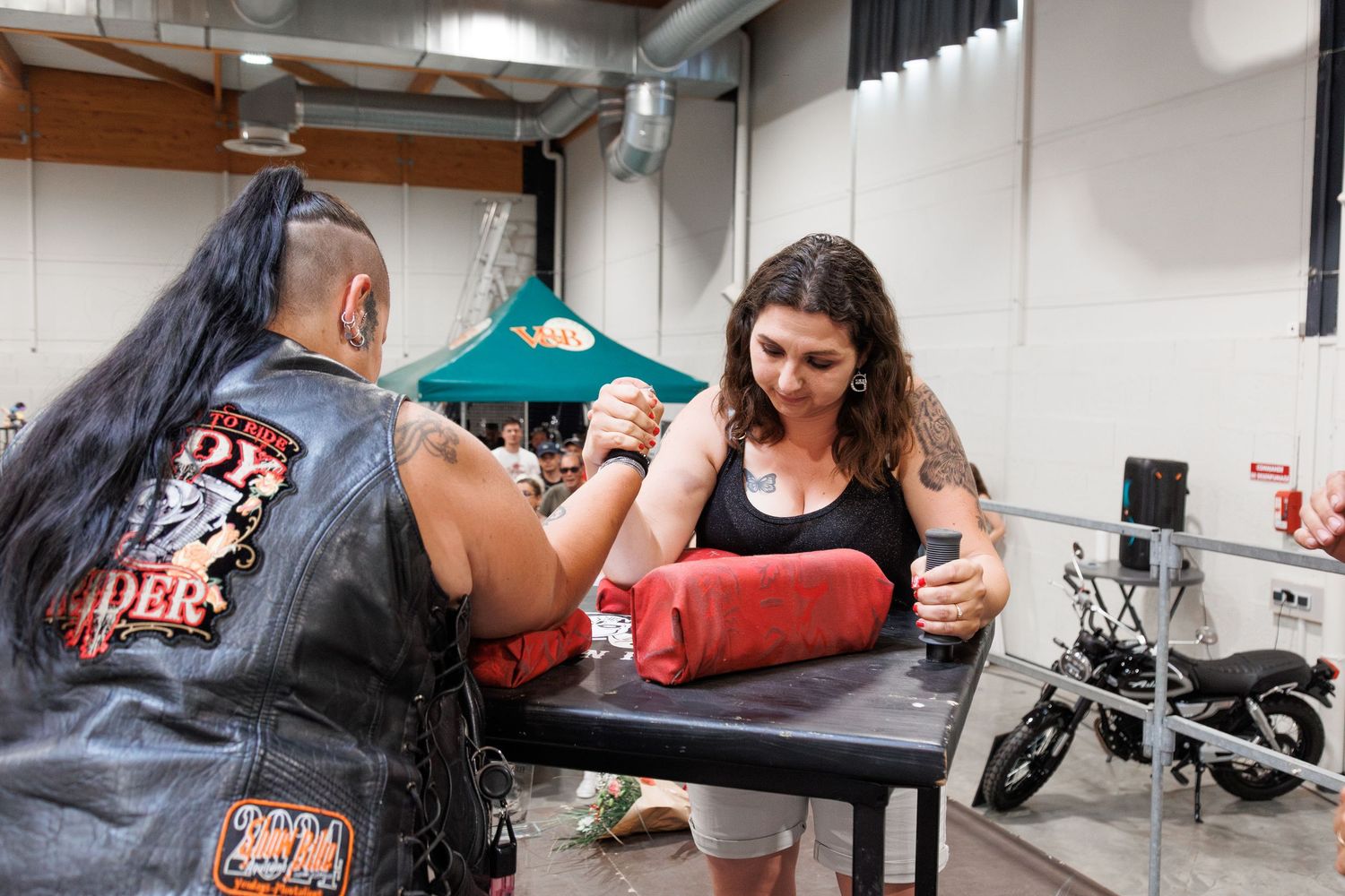 Two competitors arm wrestling on a padded table at an indoor event, with a motorcycle visible in the background.