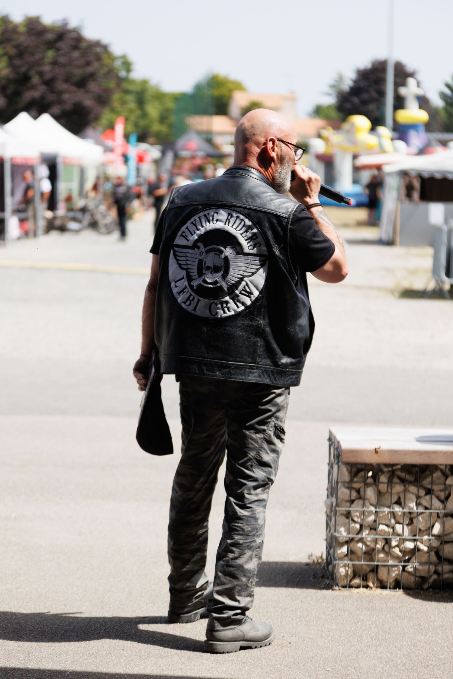 Bald man in leather biker vest with patch, talking on phone at outdoor motorcycle rally event.