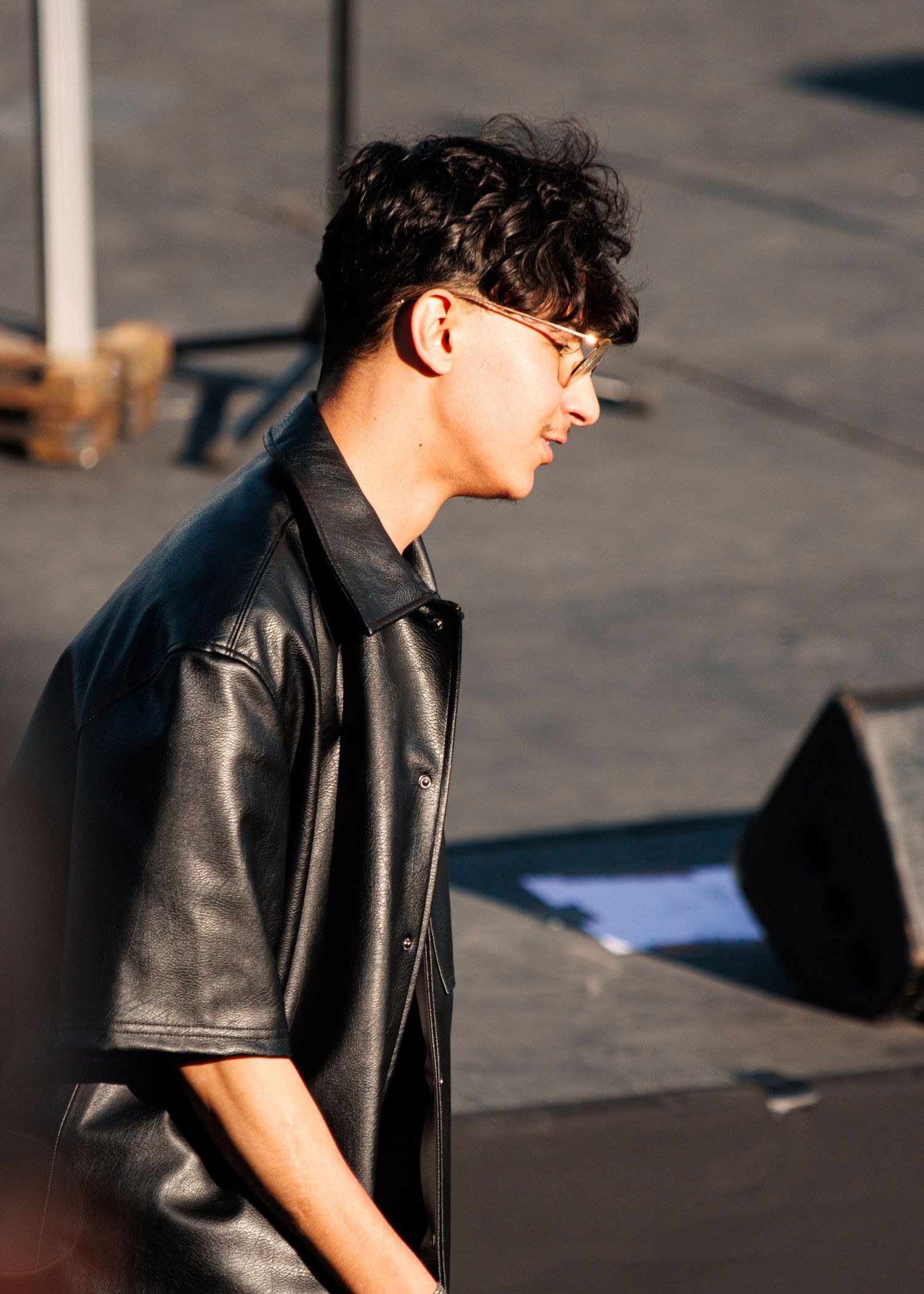 Person in sunlight wearing black leather jacket and glasses on an outdoor stage.