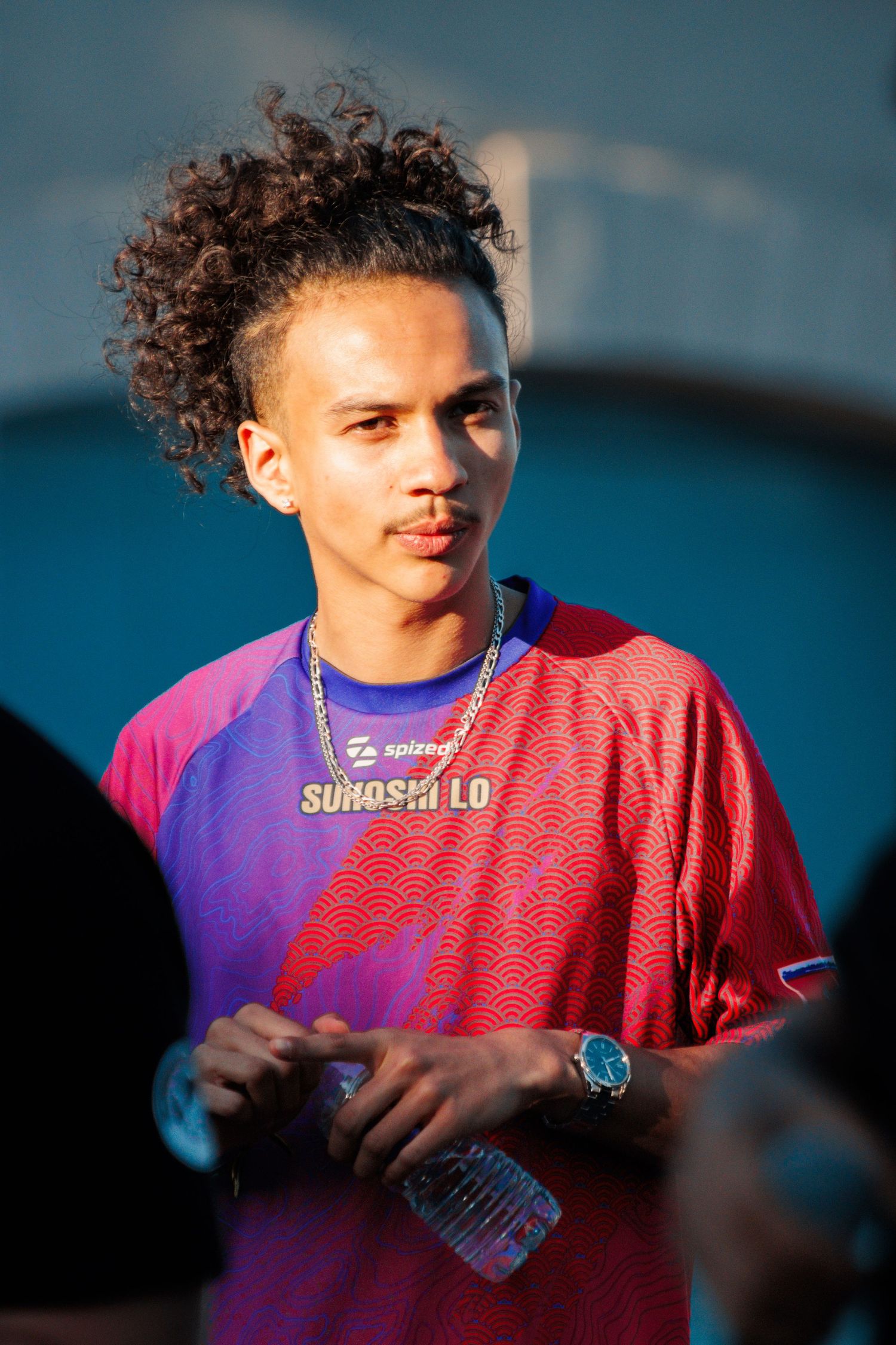 Young person in vibrant purple and pink jersey with curly hair smiling in sunlight.