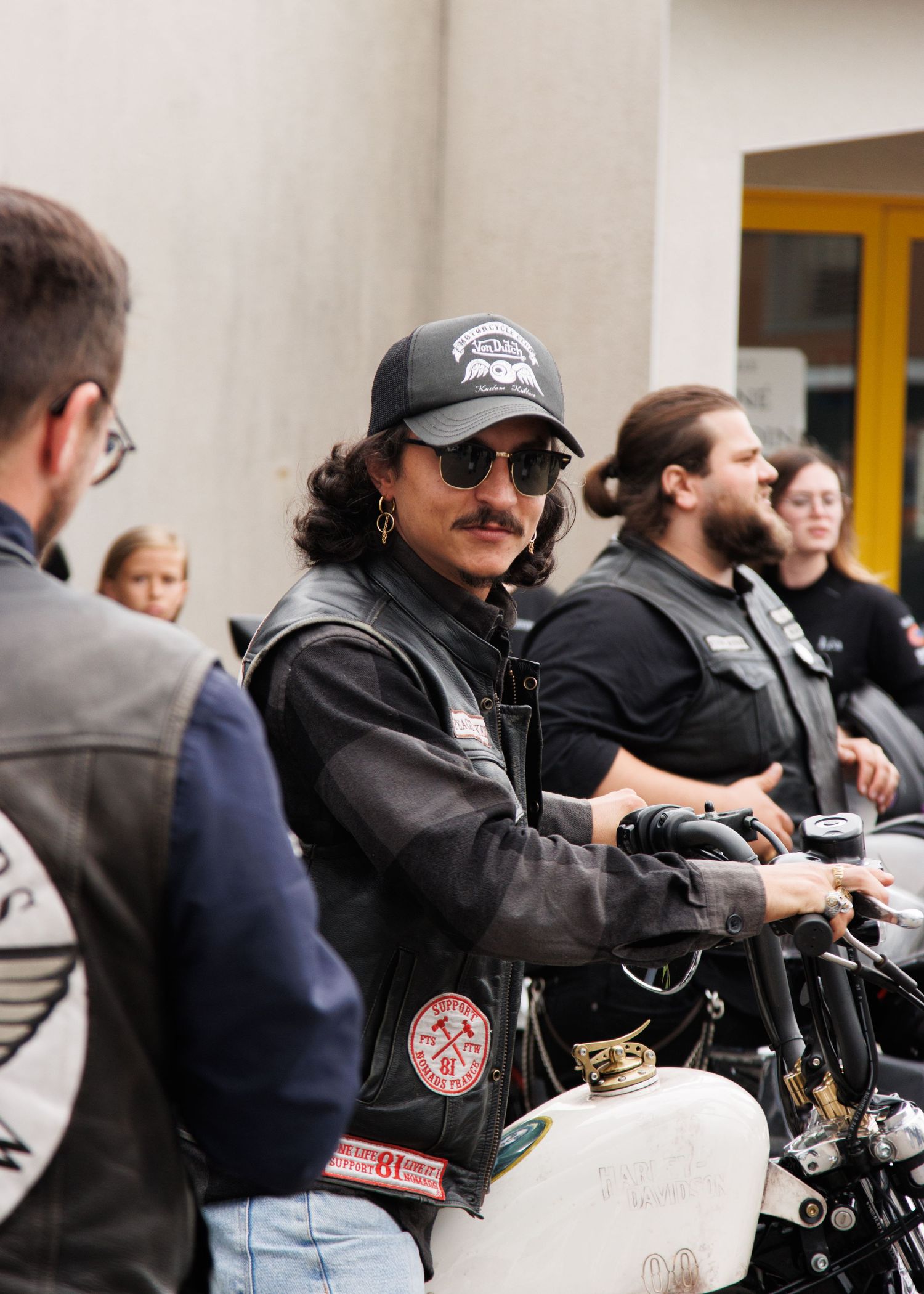 Motorcyclist in leather jacket and baseball cap stands next to bike at outdoor gathering.