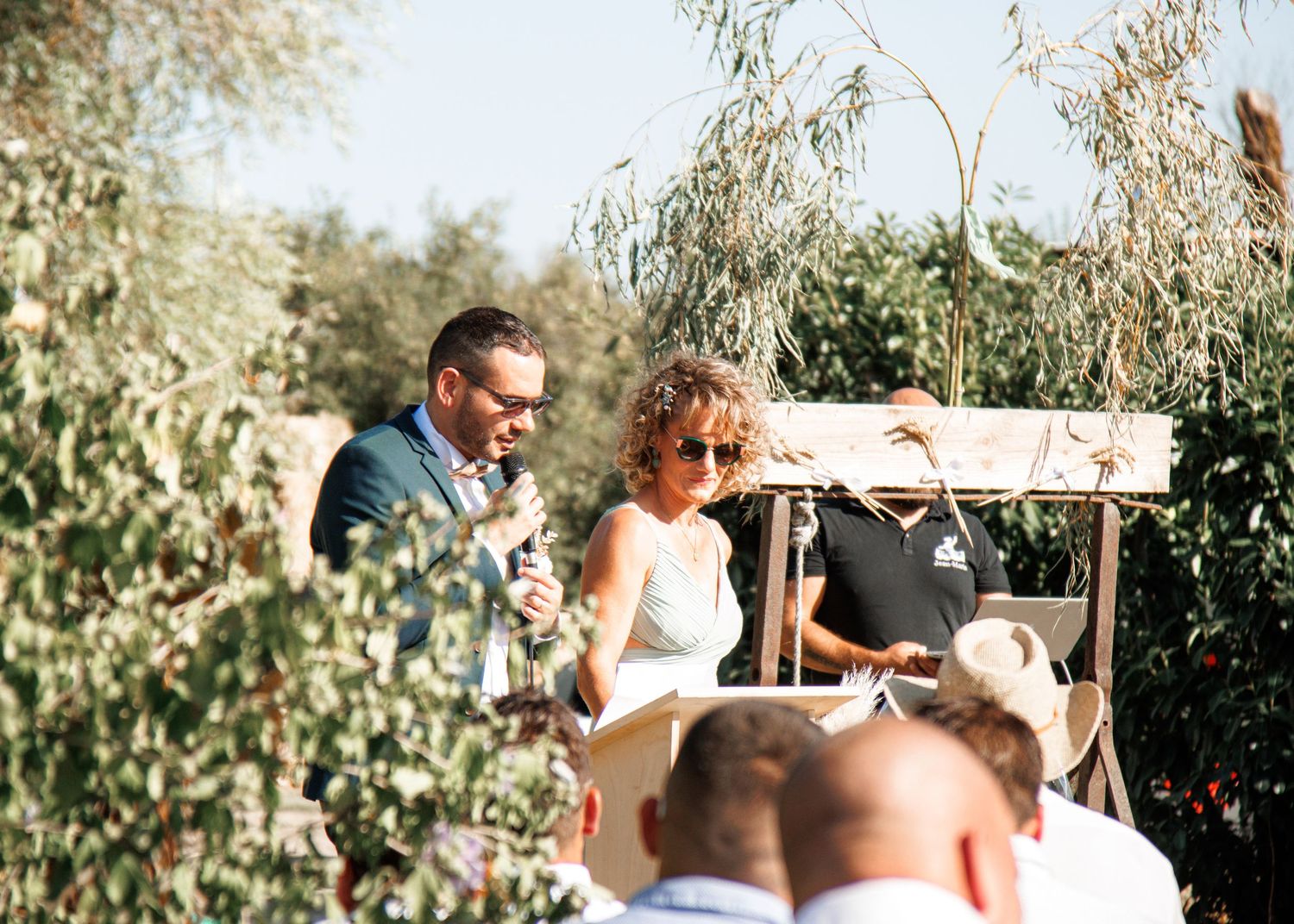 People gather outdoors at a sunny garden party with olive trees and natural decor in the background.