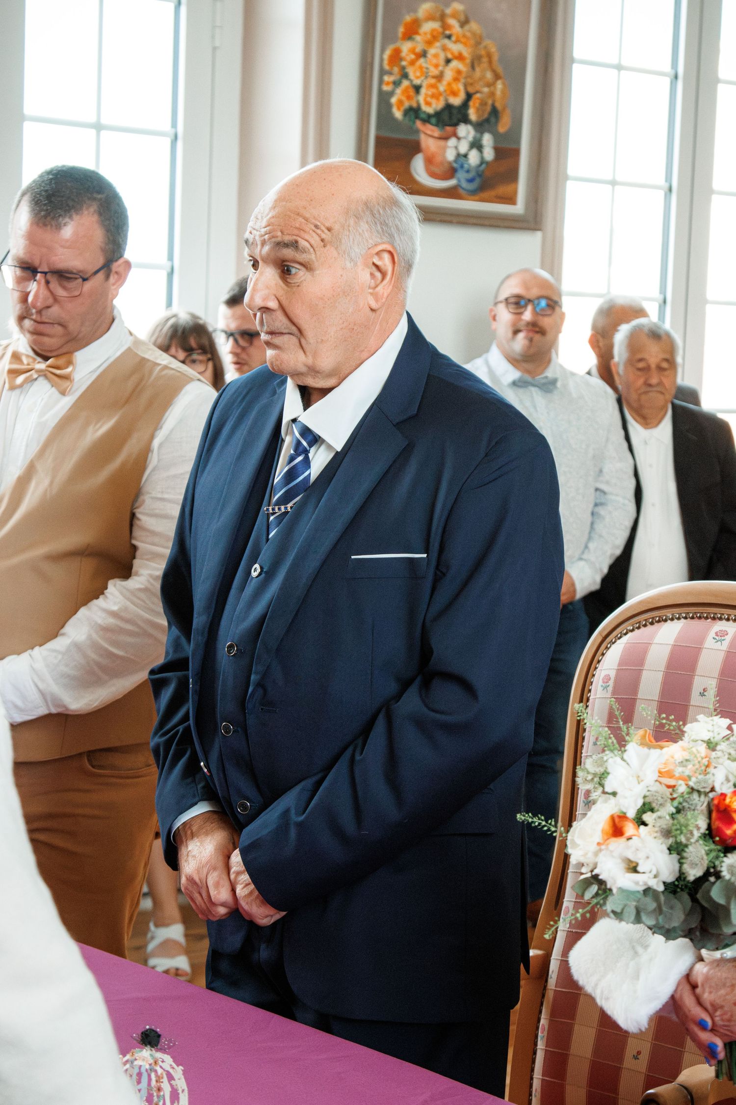 Elderly man in navy suit attending a formal wedding ceremony indoors with guests standing in background.