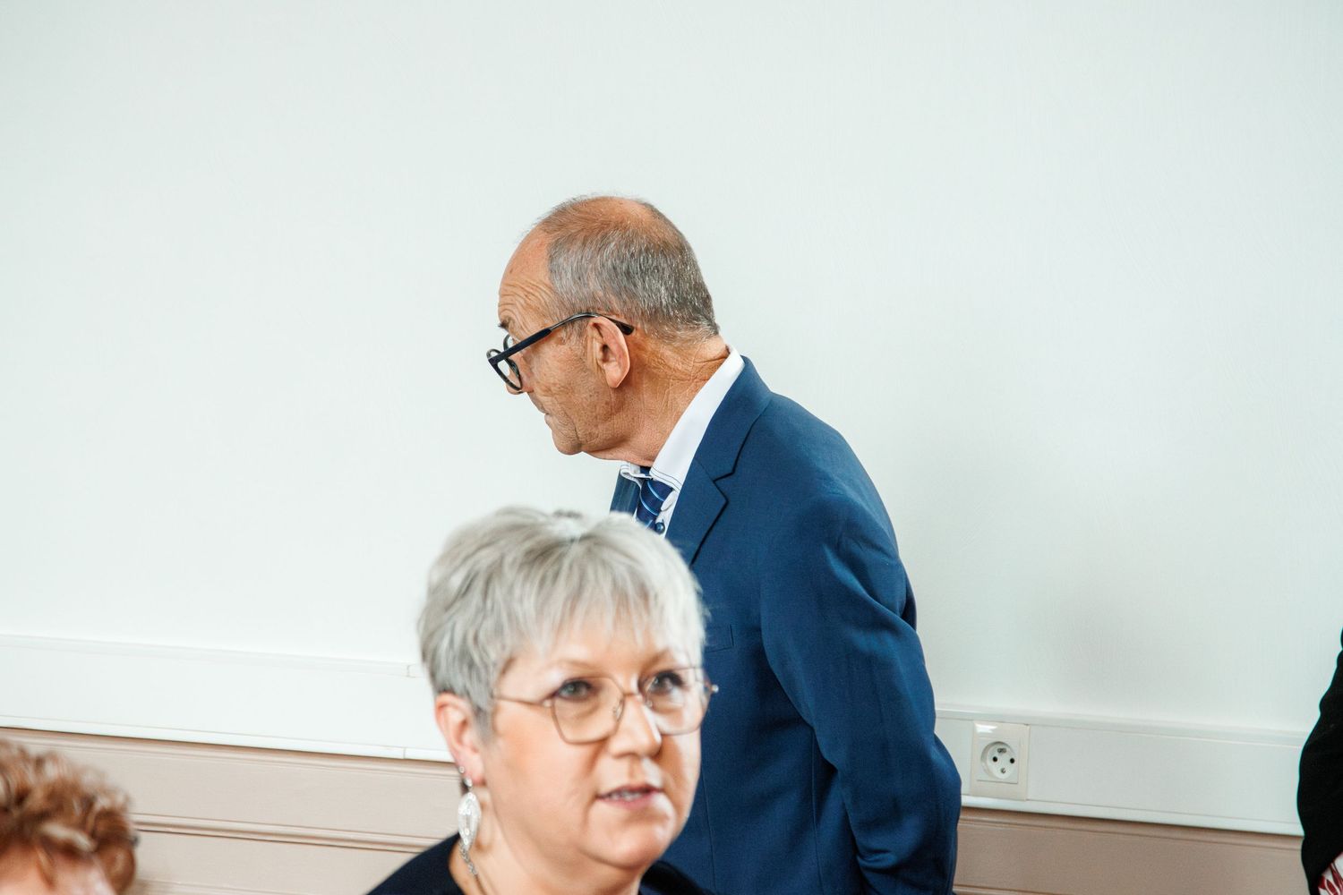 Two elderly professionals in a meeting room, man in blue suit with glasses standing behind seated woman with short gray hair.