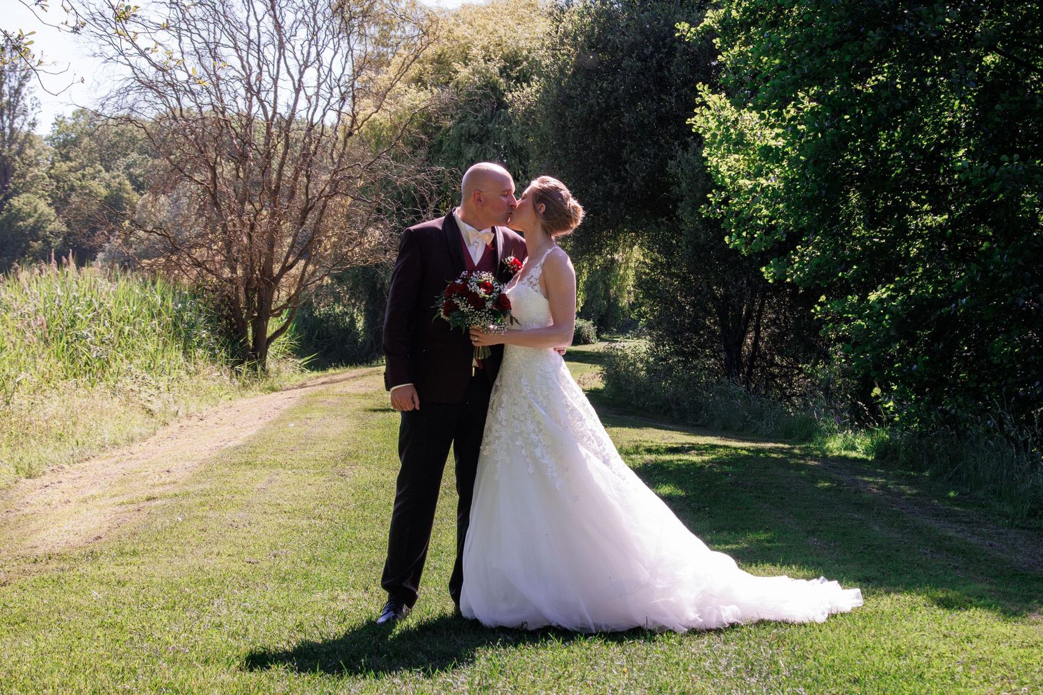 Bride and groom share a kiss on a sunny garden path, bride in lace gown holding red and white bouquet.