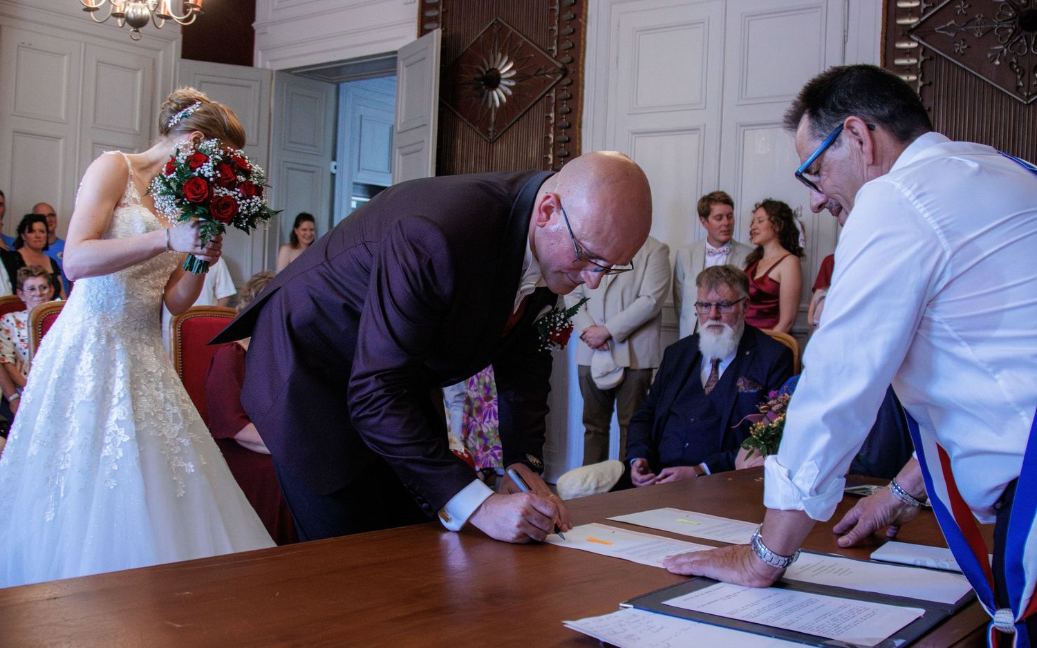 Groom signing wedding register at ceremony while bride holds red rose bouquet.