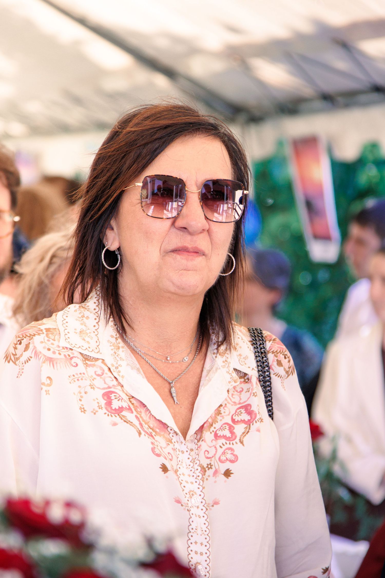 Elegant middle-aged woman wearing large square sunglasses and white embroidered blouse at an outdoor event.