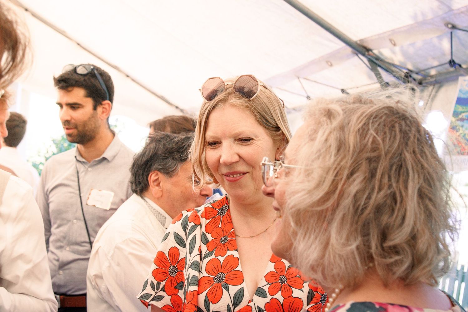 Blonde woman in red floral dress smiling and conversing with guests at outdoor wedding reception.