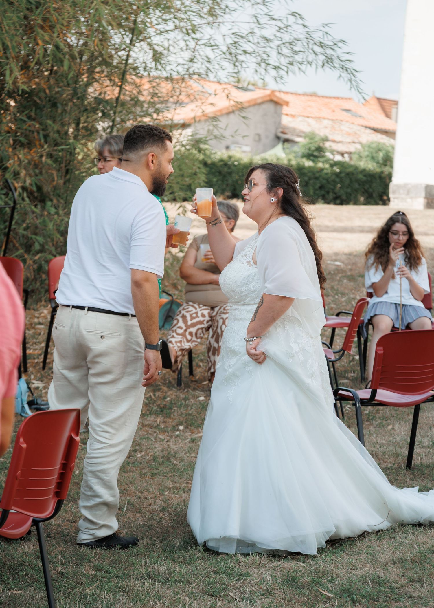 Bride in white gown toasting drinks with guest outdoors at wedding reception with red chairs.