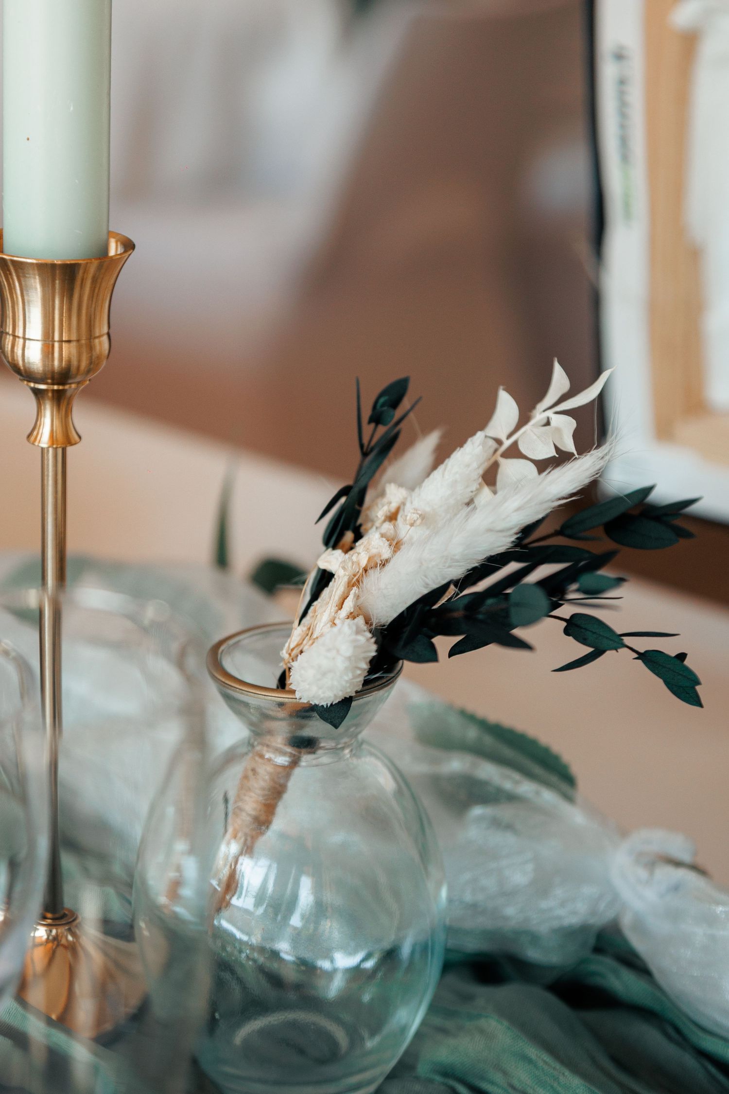 Wedding table decor with dried white flowers in glass vase beside gold candlestick with mint candle.