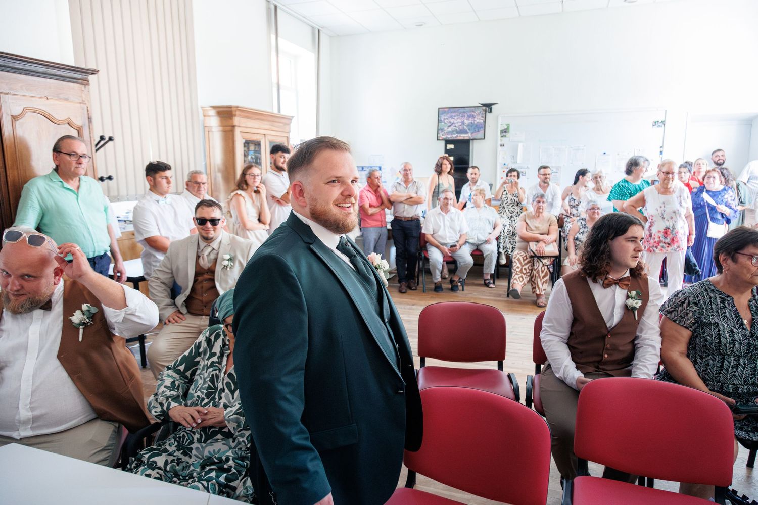 Groom in dark green suit stands smiling among seated wedding guests in a bright ceremony hall.