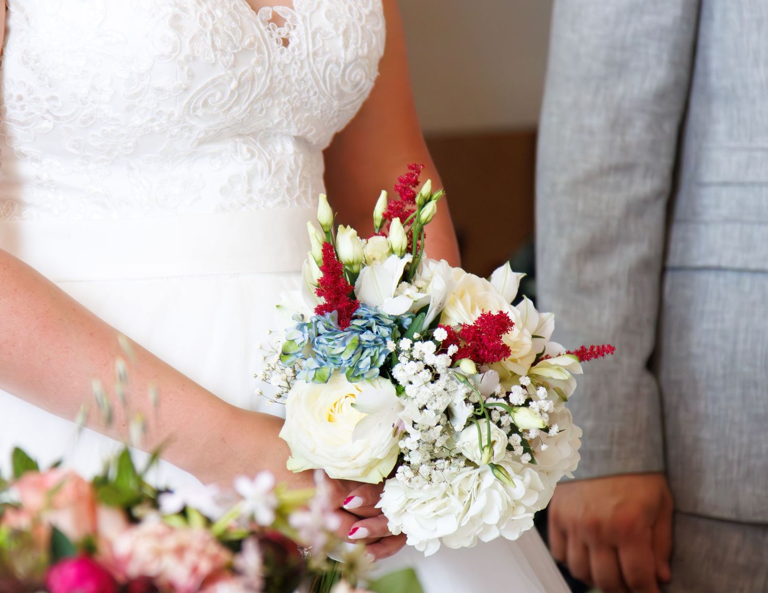 A wedding bouquet featuring white roses, red flowers, and baby's breath held against a white lace dress and gray suit.