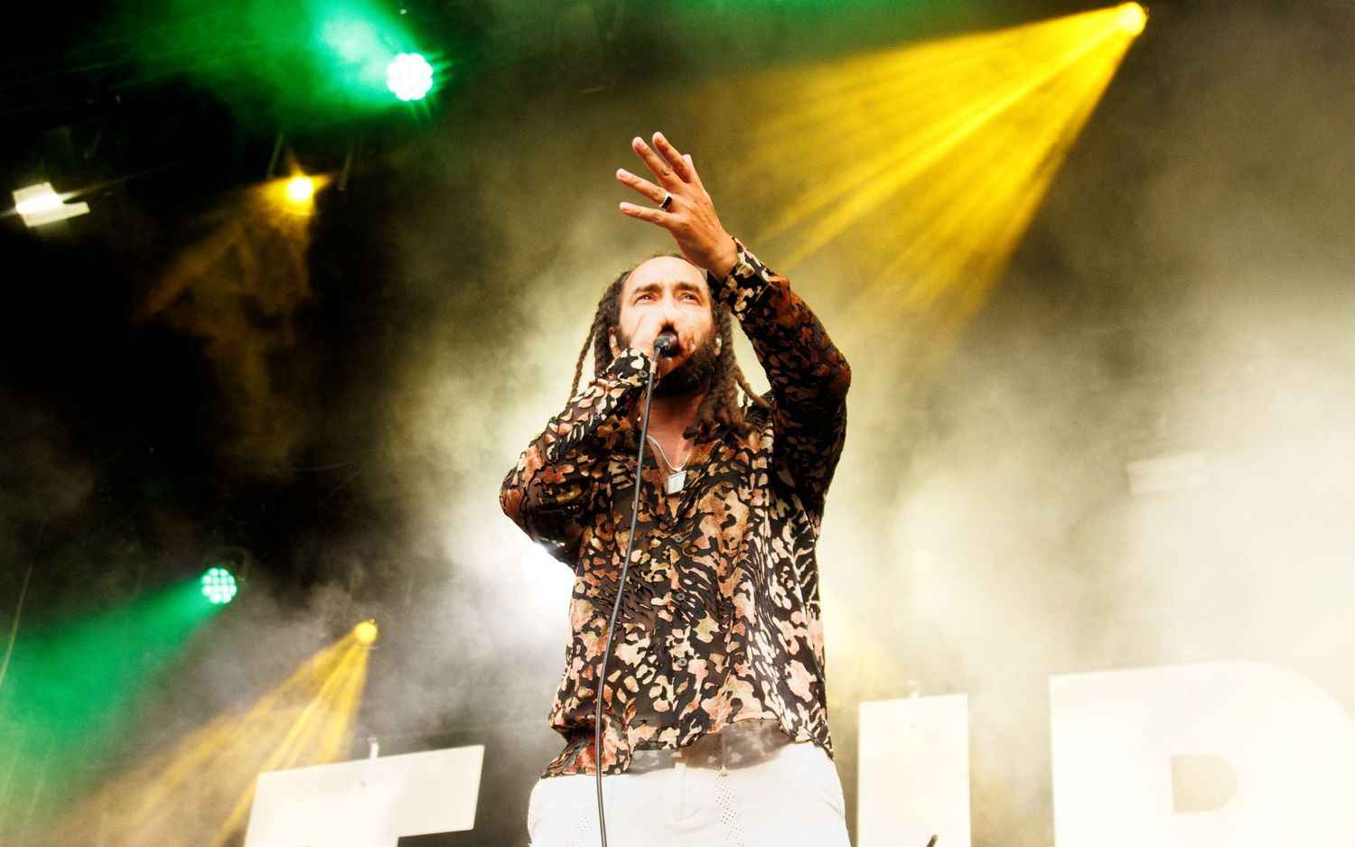 Male performer singing on stage with dramatic yellow and green stage lighting and smoke effects.