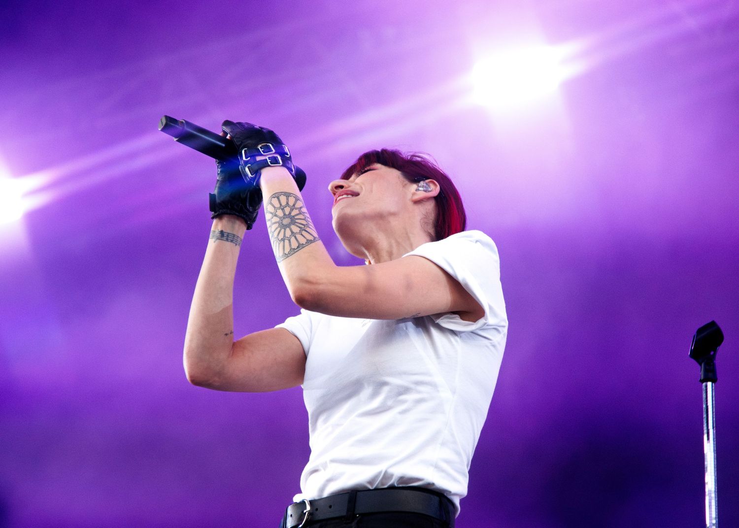 A tattooed singer performs on stage holding a microphone under purple lights wearing a white t-shirt.