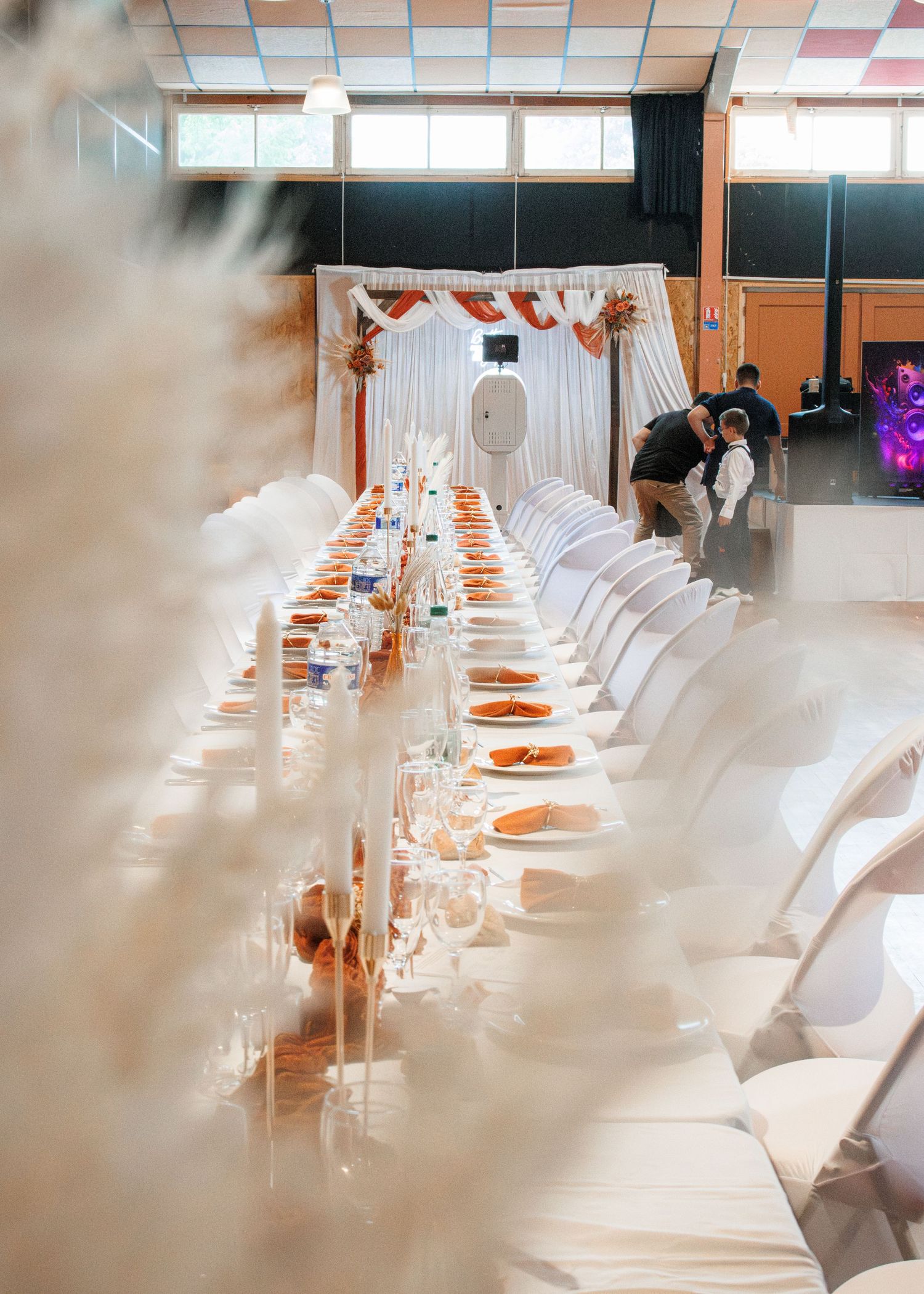 Elegant wedding reception hall with long banquet table, white chairs, orange accents, and a decorated backdrop.