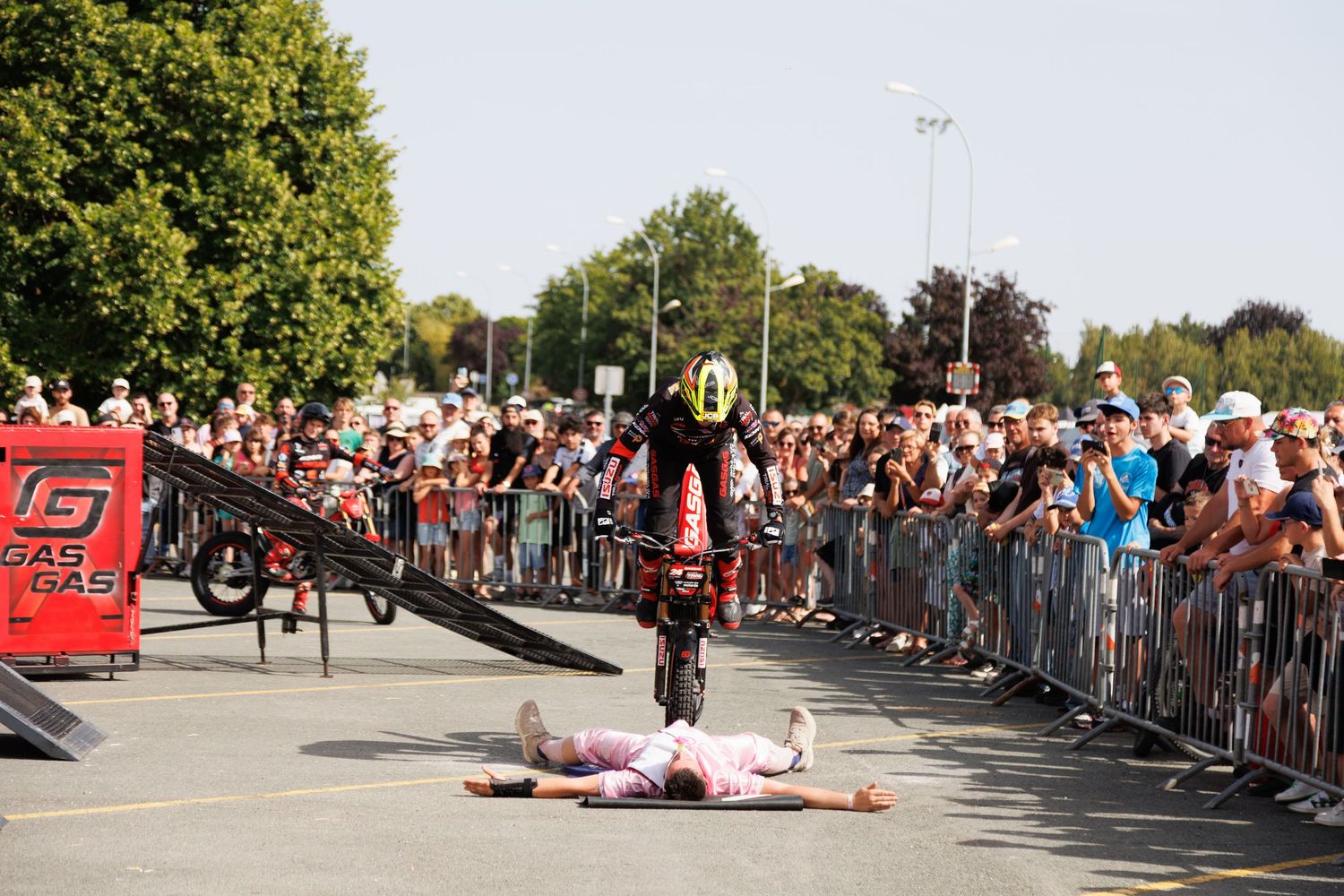 Stunt cyclist jumps over a person lying on the ground at an outdoor show, with a large crowd watching behind metal barriers.