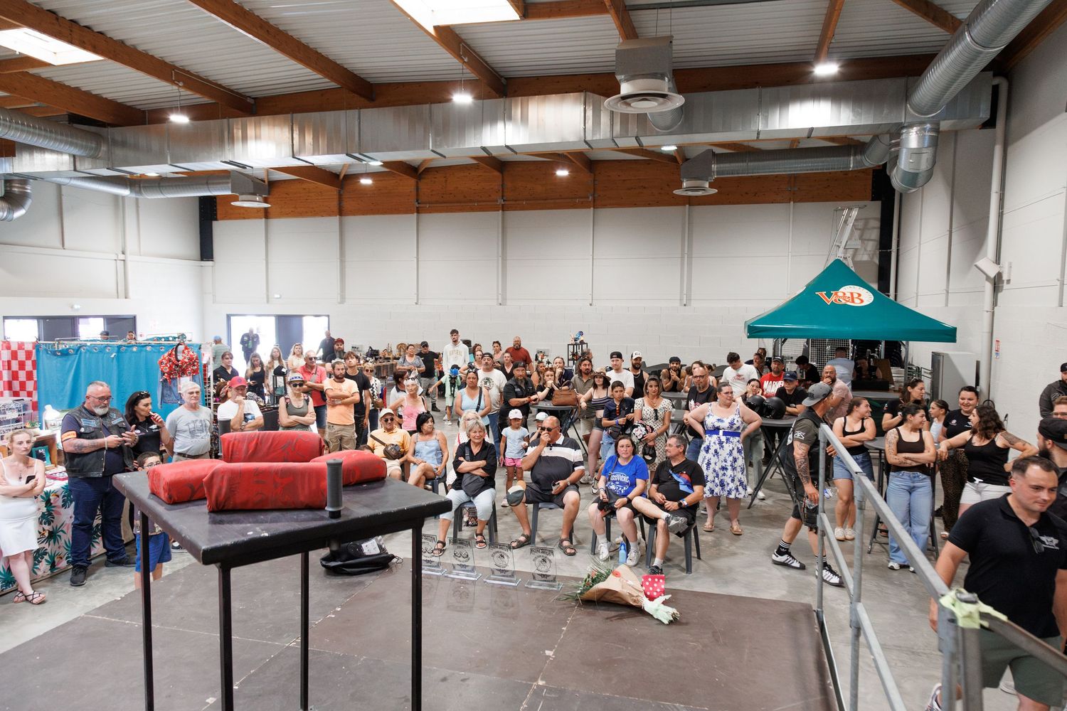 Large crowd gathered inside an industrial event hall with a stage, green tent, and red couch during an indoor gathering.