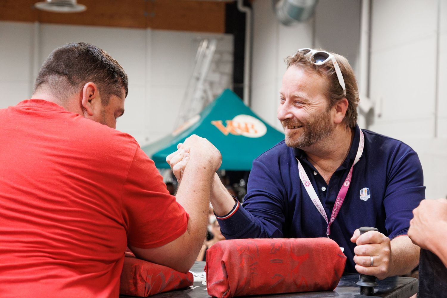 Two men arm wrestling at a table outdoors, smiling and competing in a friendly match.