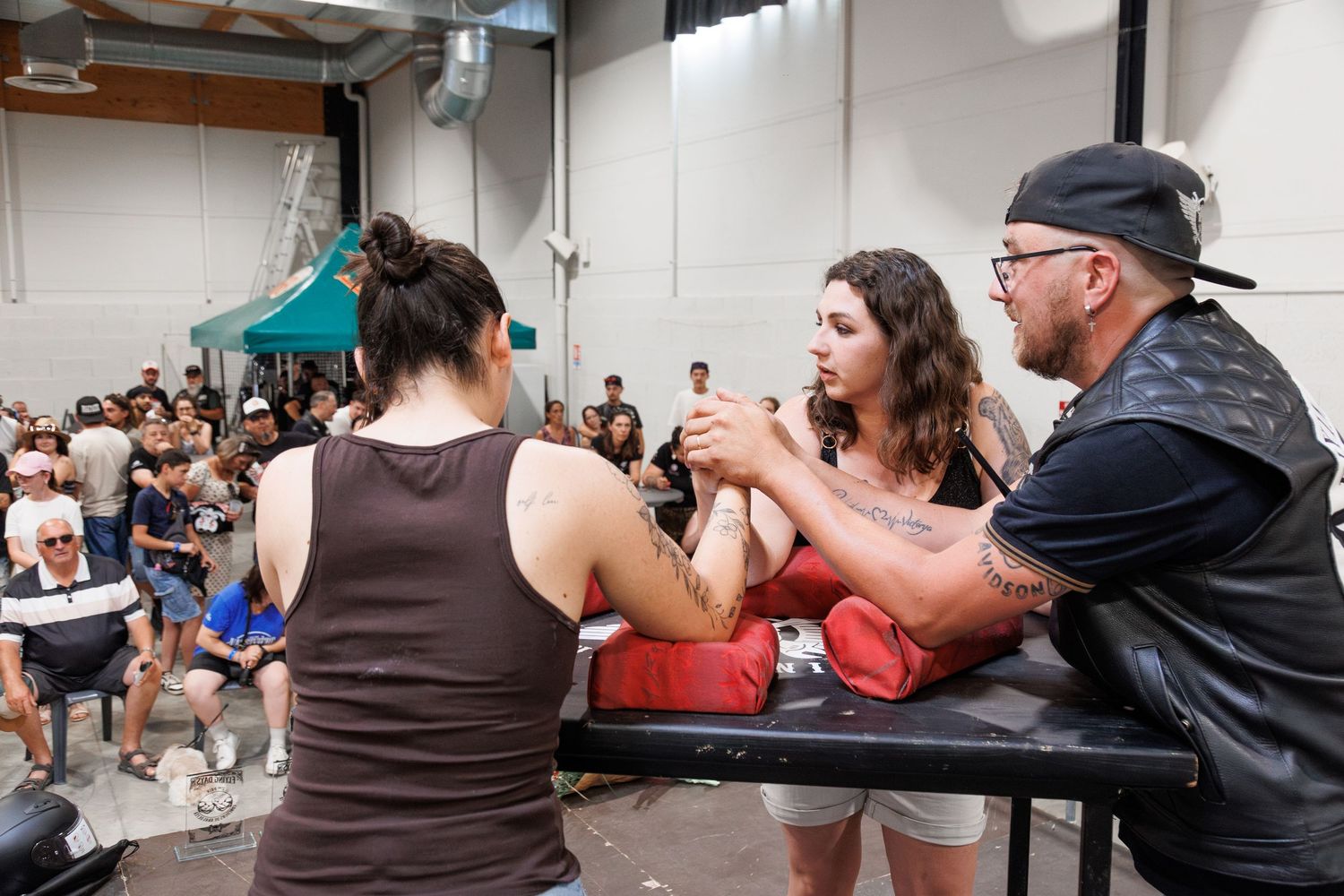 Two competitors arm wrestling at a table during an indoor arm wrestling competition with a crowd watching in the background.