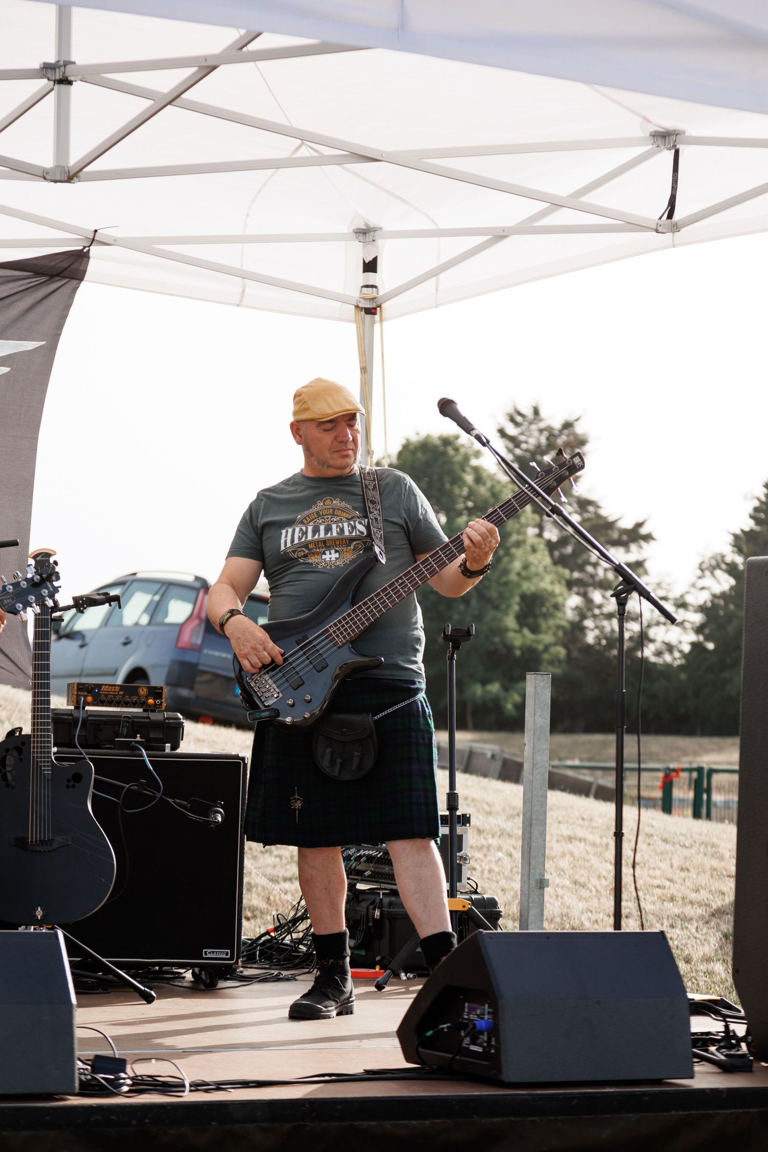 Male guitarist in kilt and Hellfest shirt performs on outdoor stage under white canopy tent.