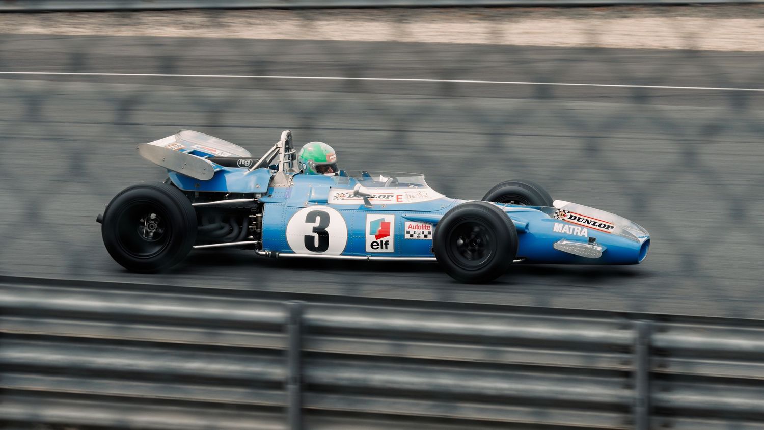 Blue and white formula racing car speeds along track during competitive motorsport event.
