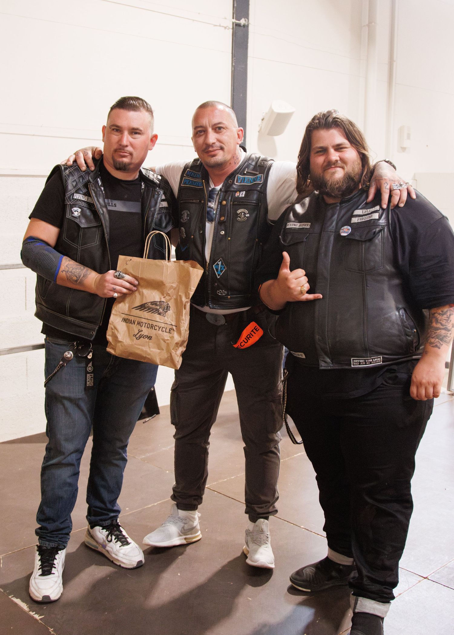 Three bikers in leather vests pose together holding a brown paper bag in an indoor setting.