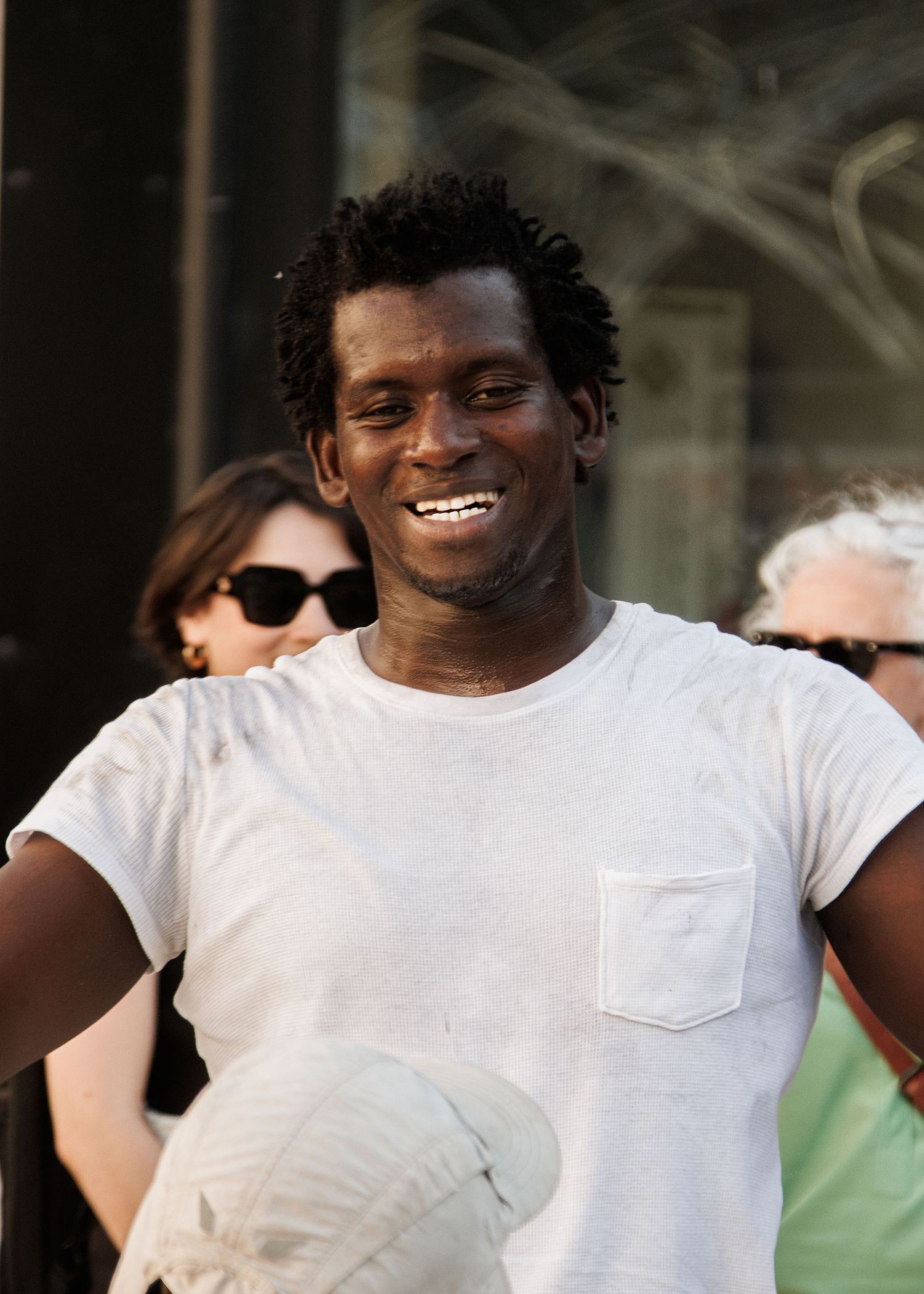 Performer in white t-shirt smiles warmly at outdoor event with crowd in background.