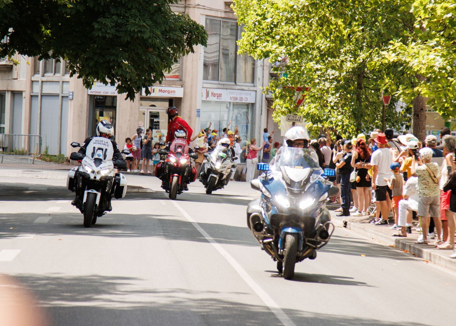 Police motorcycles lead a professional cycling race through a tree-lined city street as spectators watch from sidewalks.