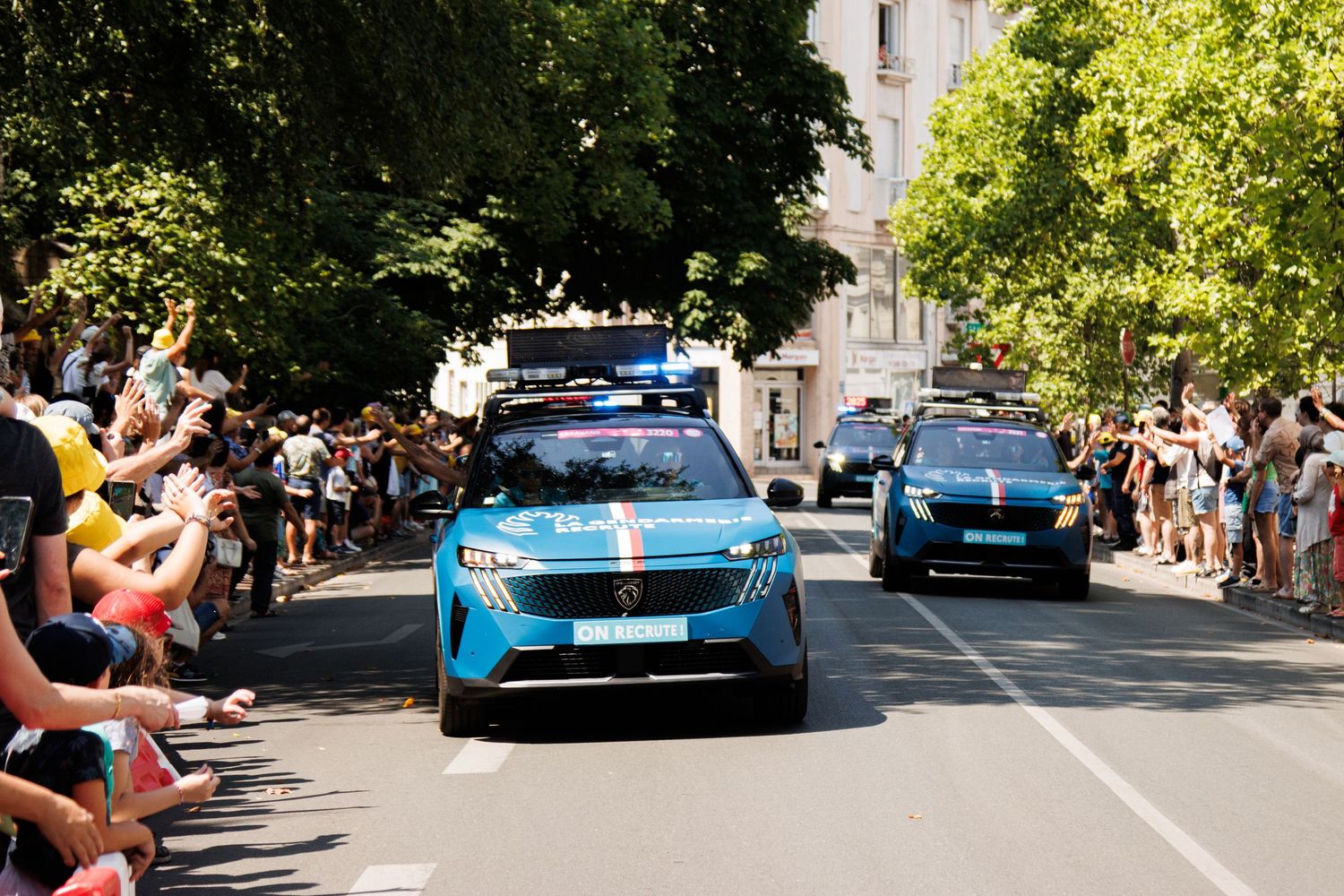 Sports cars parade down a tree-lined street past crowds of onlooking spectators.