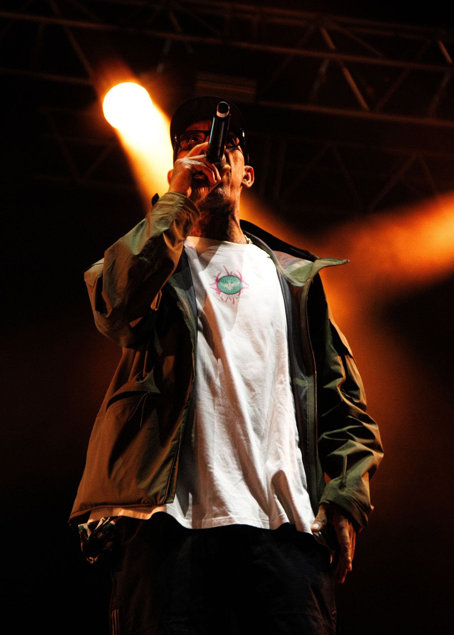 Stage performer in white t-shirt and dark jacket illuminated by dramatic orange lighting during concert performance.