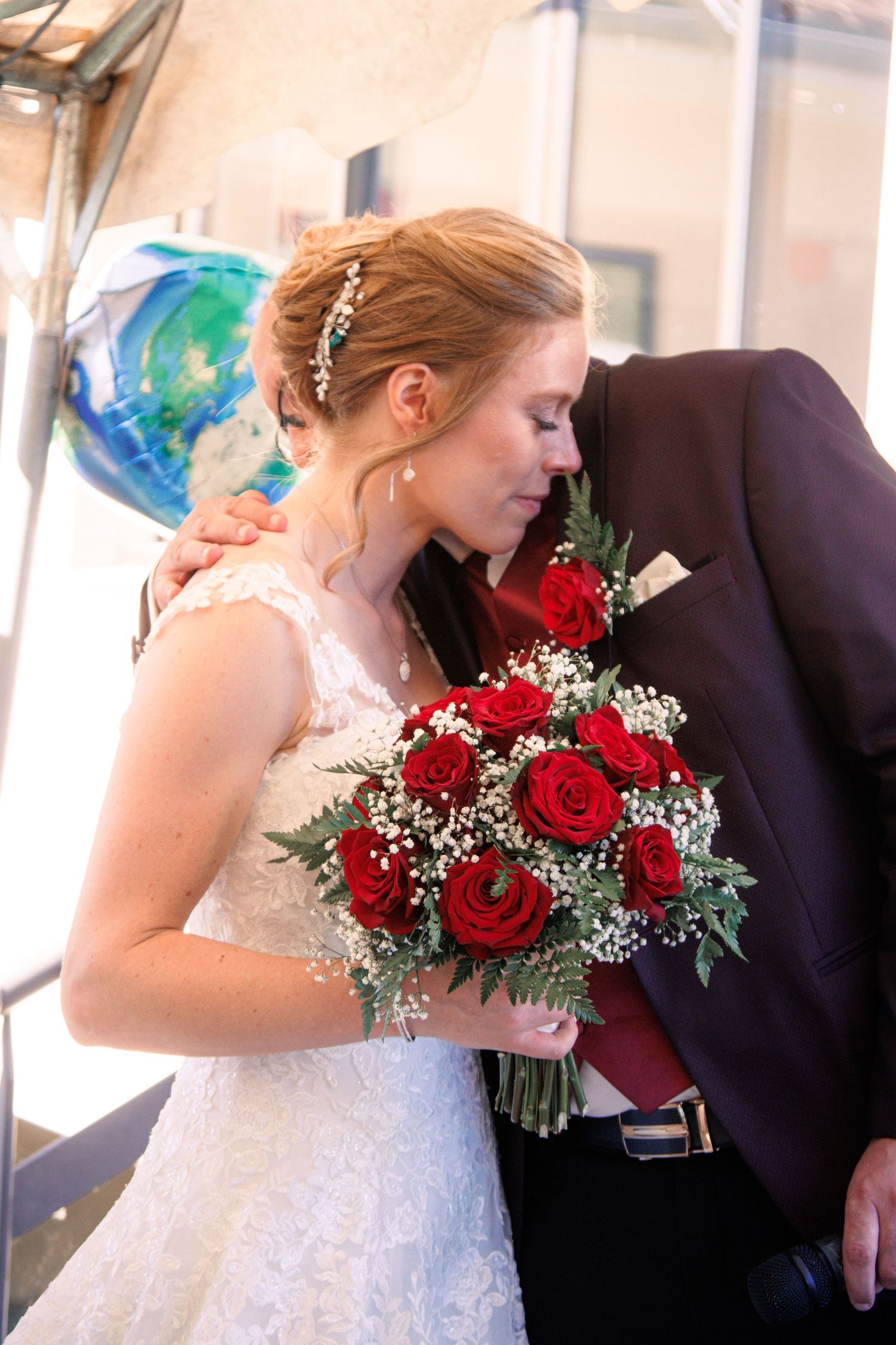 Bride in lace gown embraces groom while holding stunning red rose bouquet with baby's breath at wedding reception.