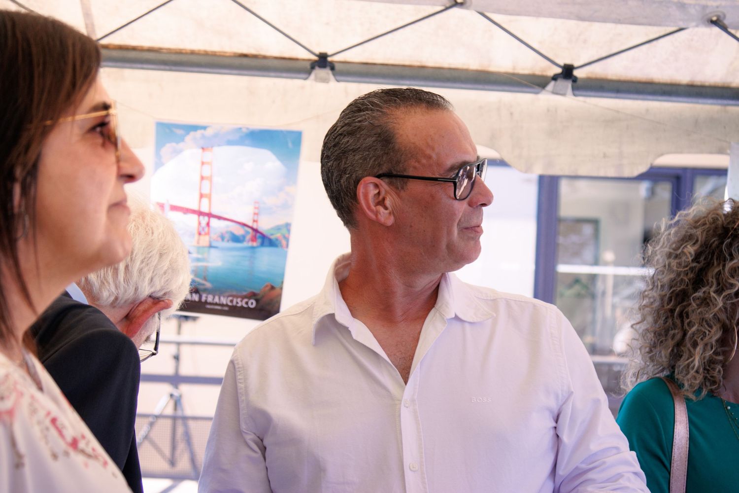 Man in white shirt and woman converse under a tent with a San Francisco Golden Gate Bridge poster behind.