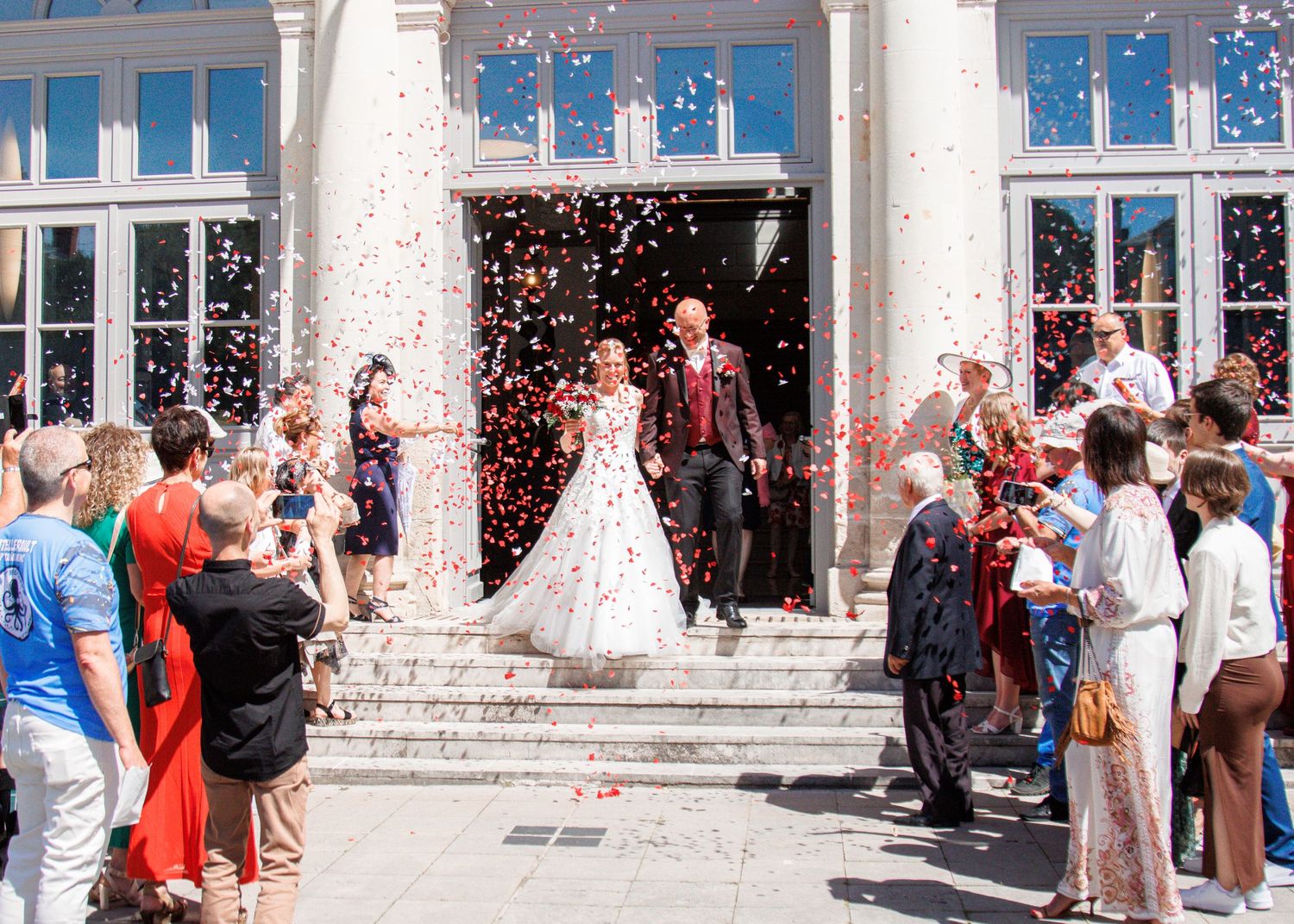 Bride and groom exit wedding venue as guests throw colorful confetti on sunny day.