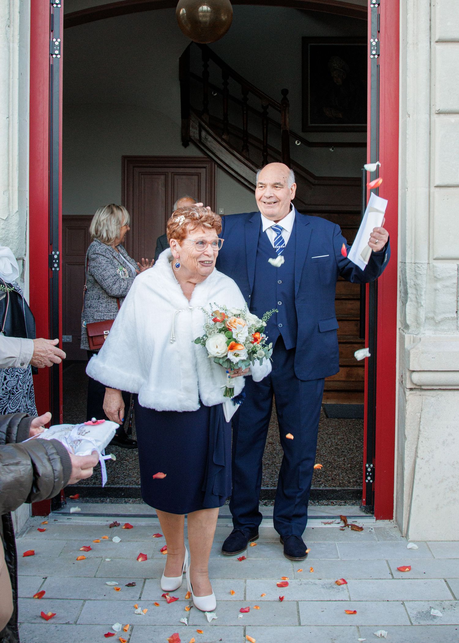 Elderly couple exits wedding venue smiling, bride holds bouquet, groom raises document, confetti on ground.