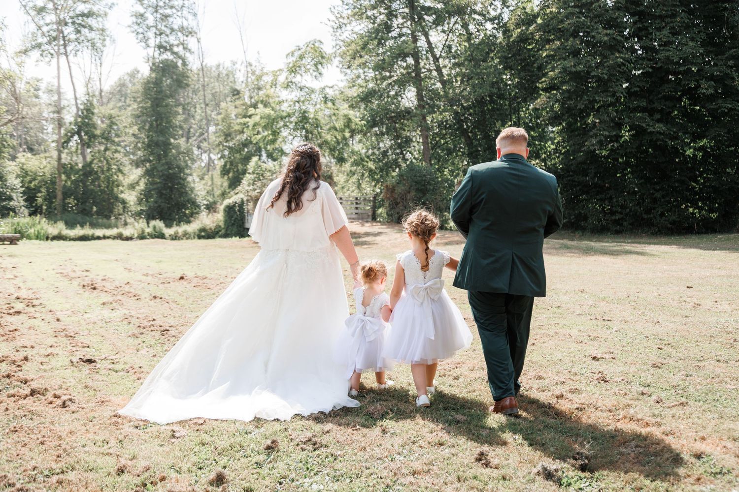 Bride and groom walking hand-in-hand with flower girl in white dress through lush green park on wedding day.