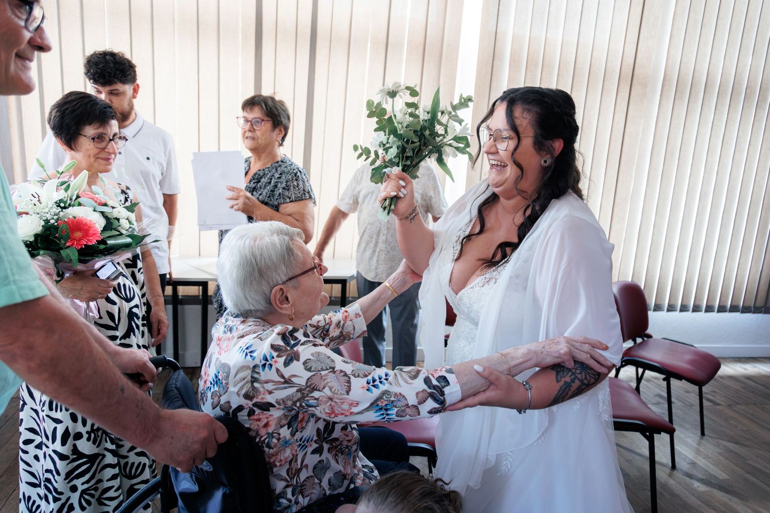 Tattooed bride in white dress laughing and greeting elderly seated guest at intimate wedding reception.