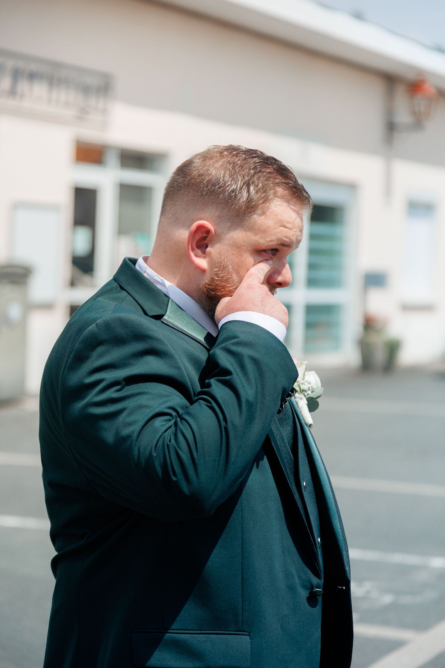 Emotional groom in dark green suit wiping tears outside on wedding day with boutonniere on lapel.