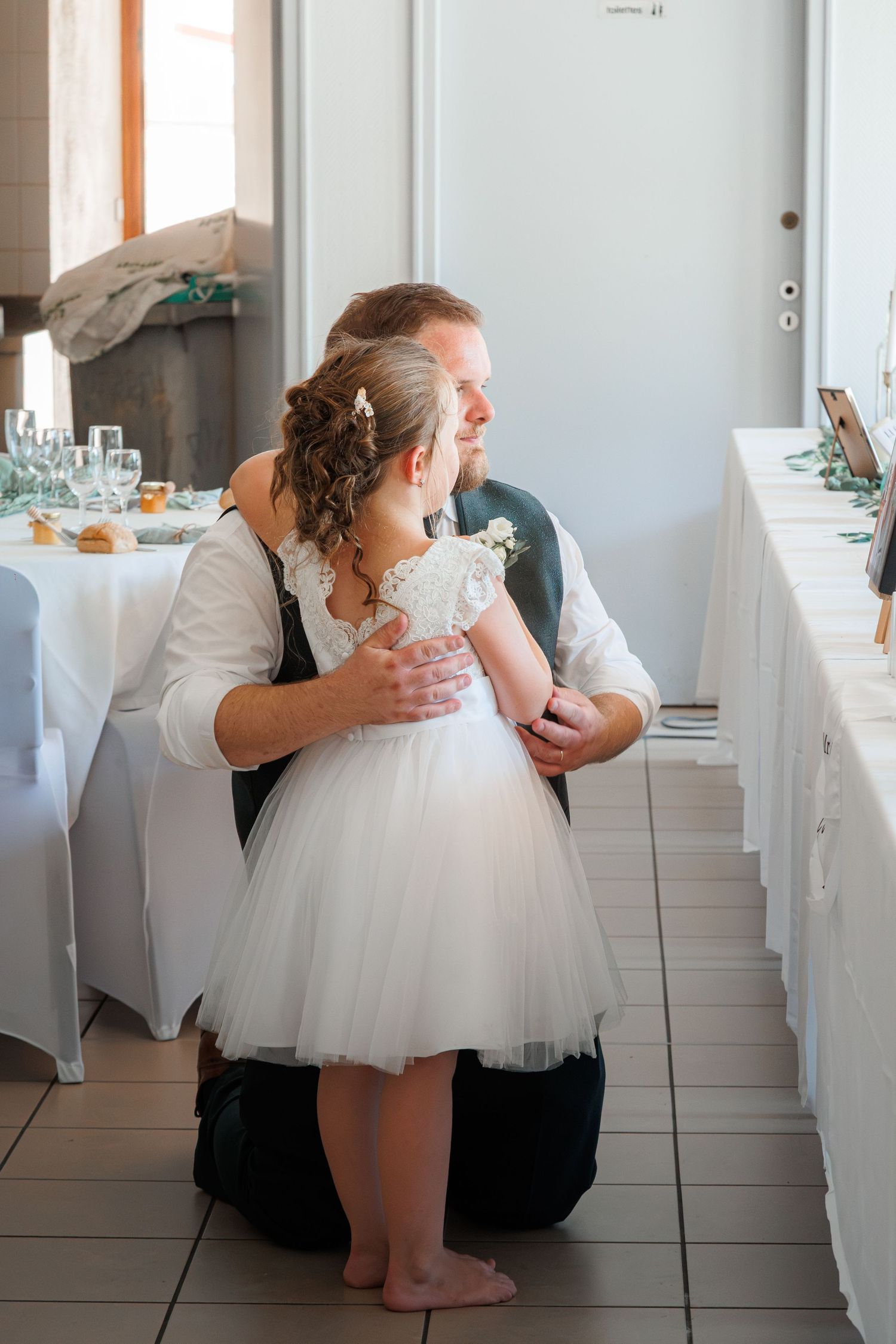 Groom in suit kneeling to embrace young flower girl in white tutu dress at wedding reception.