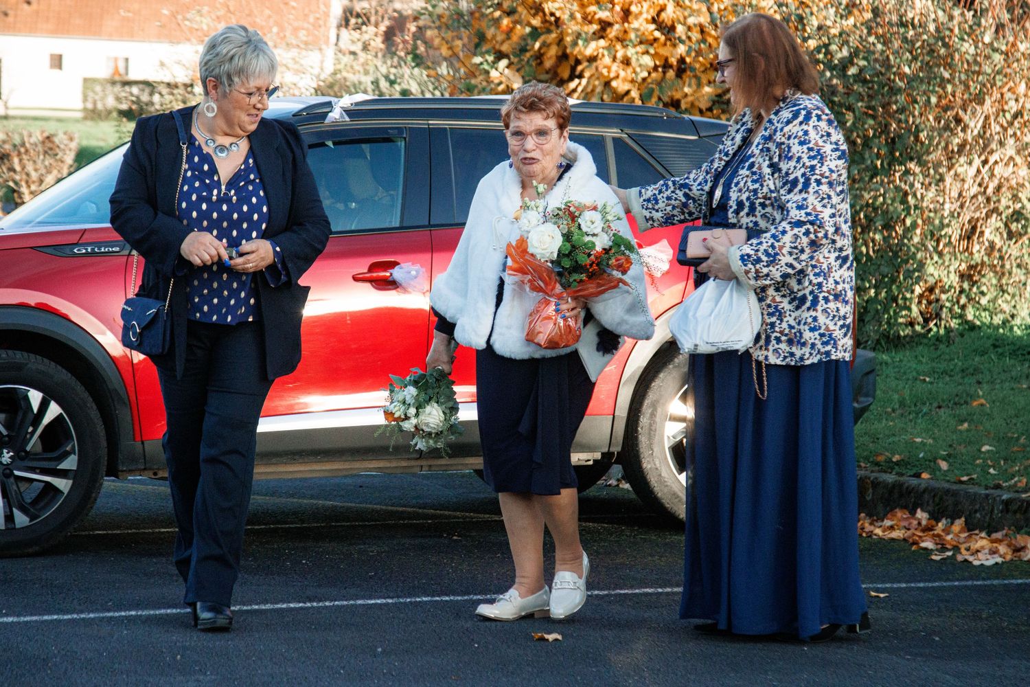 Three women gathered outdoors near a red car, with one holding a bouquet of flowers on an autumn day.