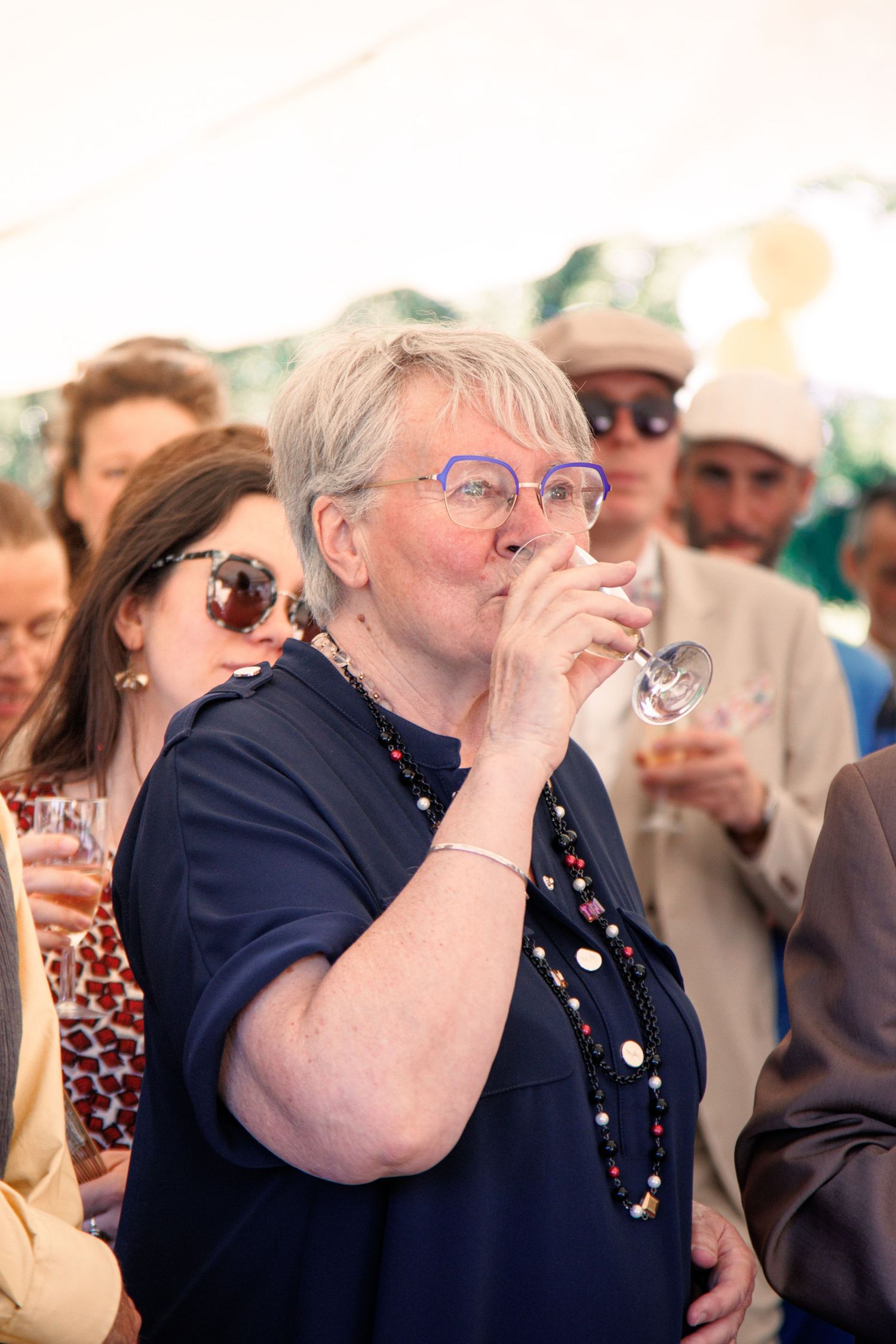 Silver-haired woman in navy dress with purple glasses drinking from champagne flute at crowded wedding reception.